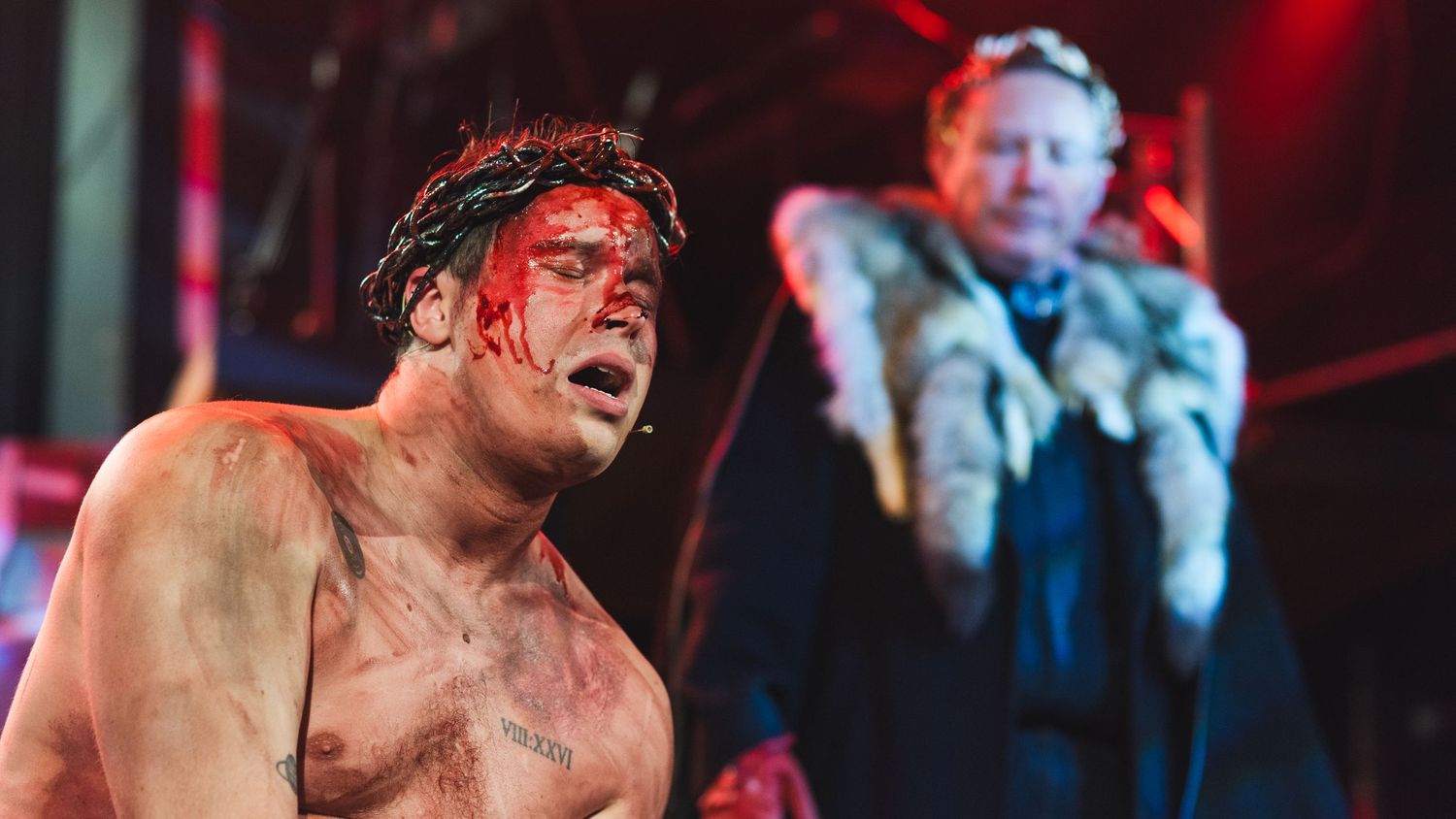 A photo from a production of Jesus Christ Superstar. The actor playing Jesus is featured, bleeding from his scalp, as Pilate looks on.
