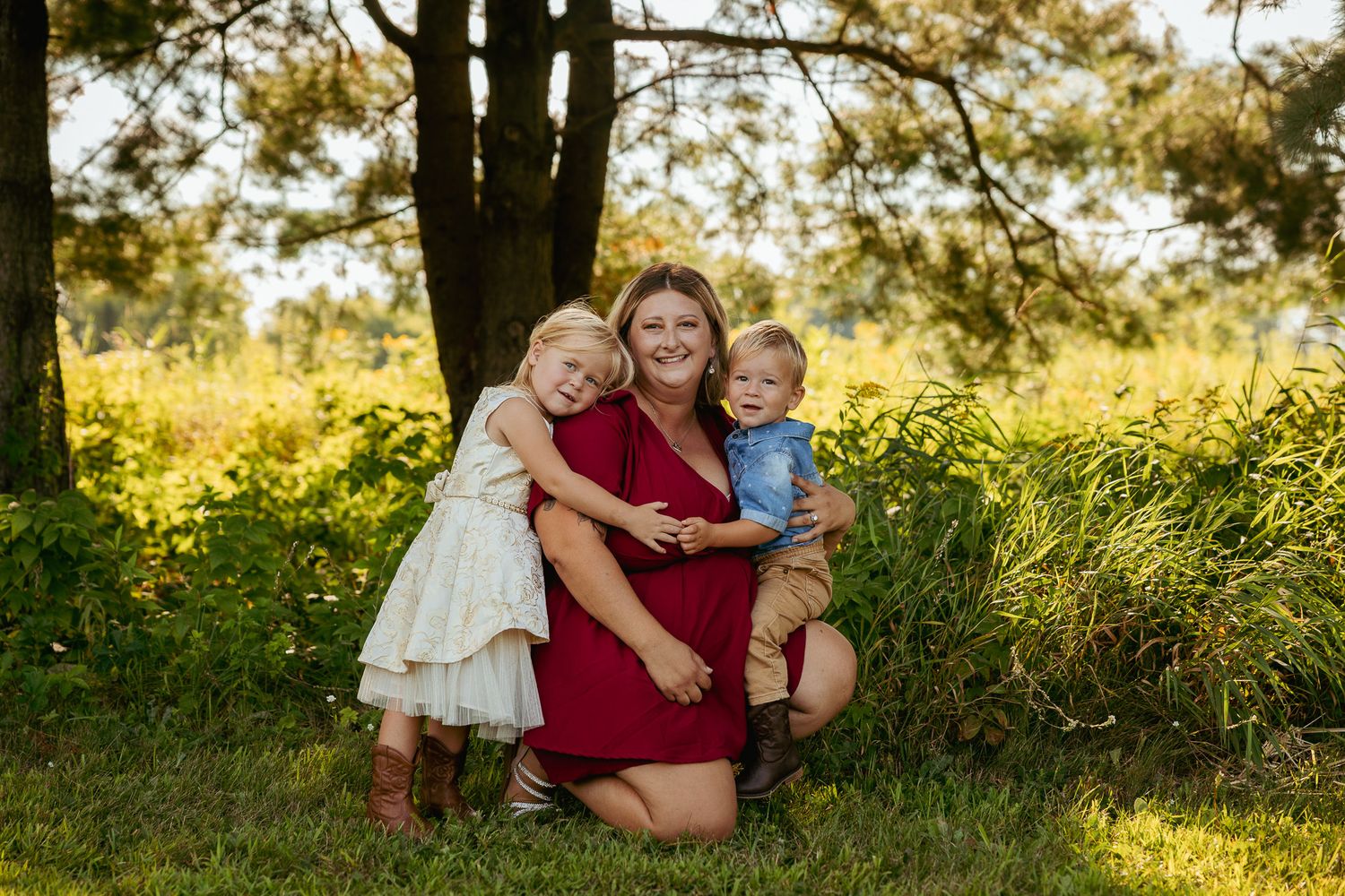 A family sits together in a grassy field during golden hour as sunlight filters through trees in the background.