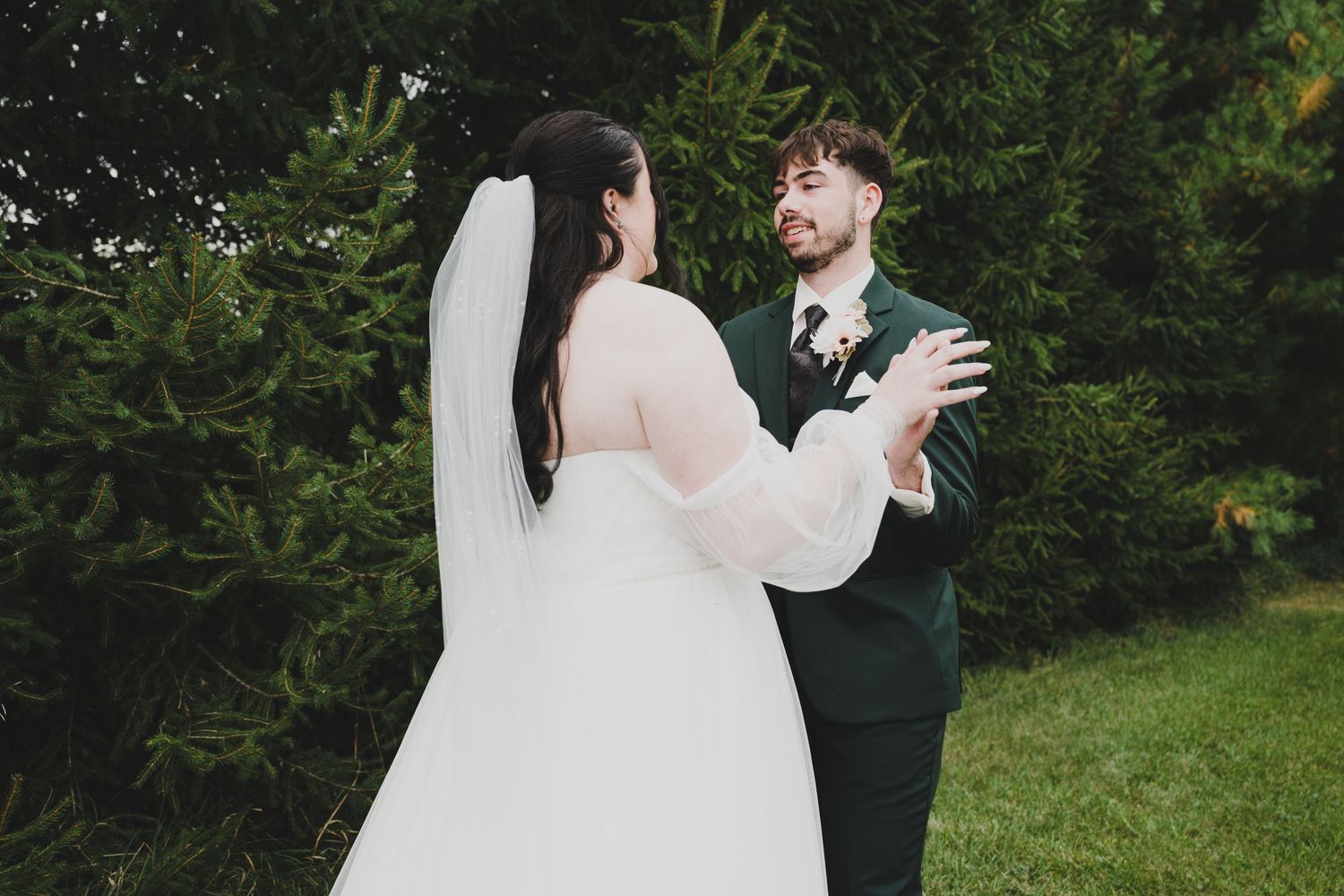 Bride in white gown and veil shares a tender first look moment with groom in dark green suit outdoors.