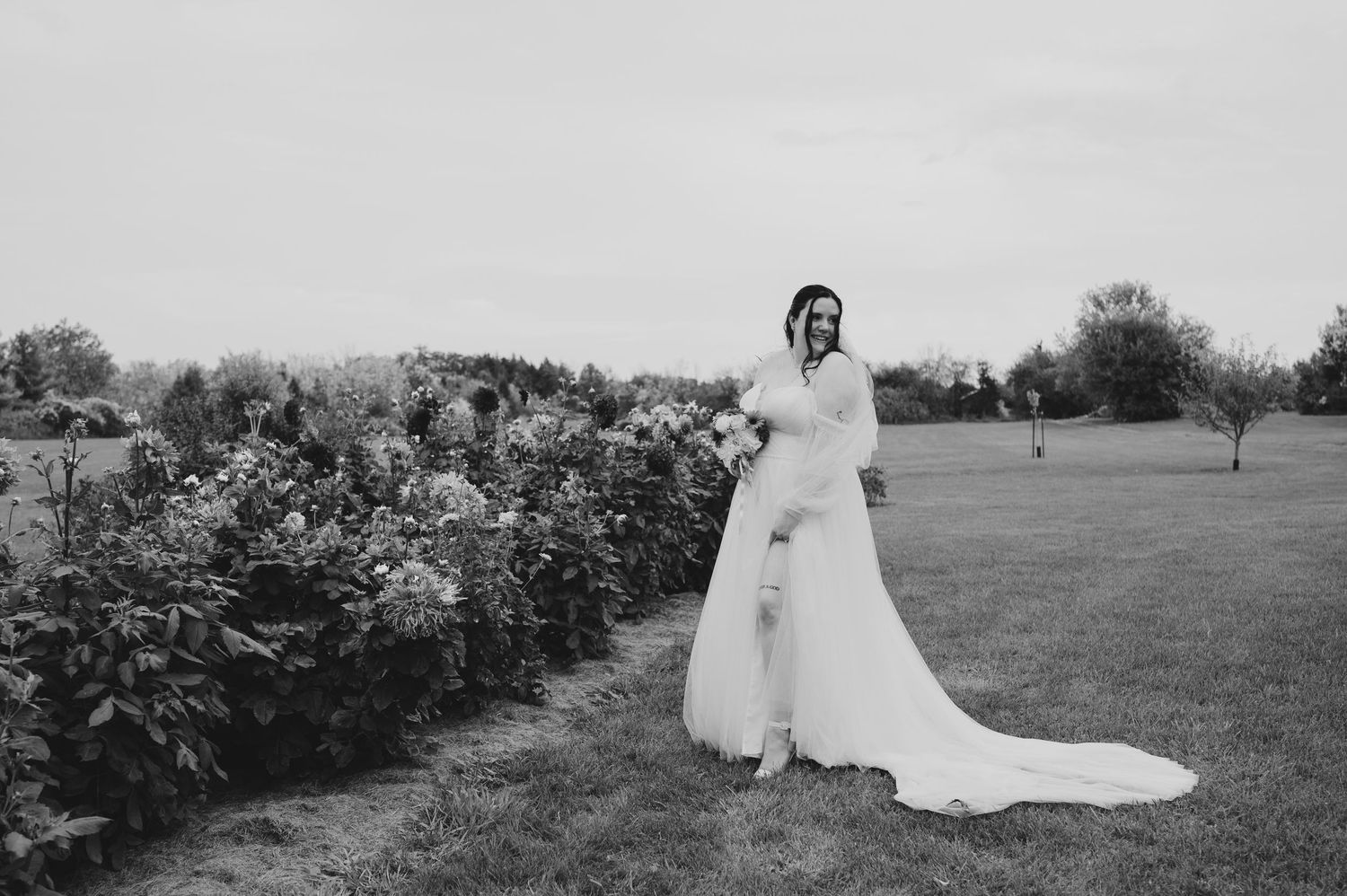 A bride in a flowing white gown poses in a garden setting with blooming flowers and trees in the background.