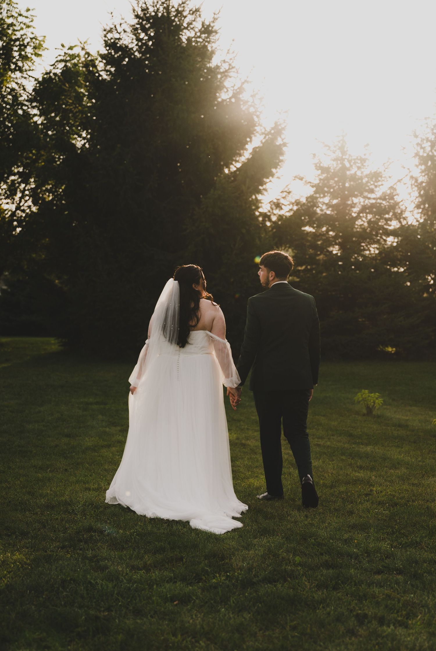 Newlywed couple walks together through a sunlit meadow at golden hour, with the bride's flowing white veil trailing behind.