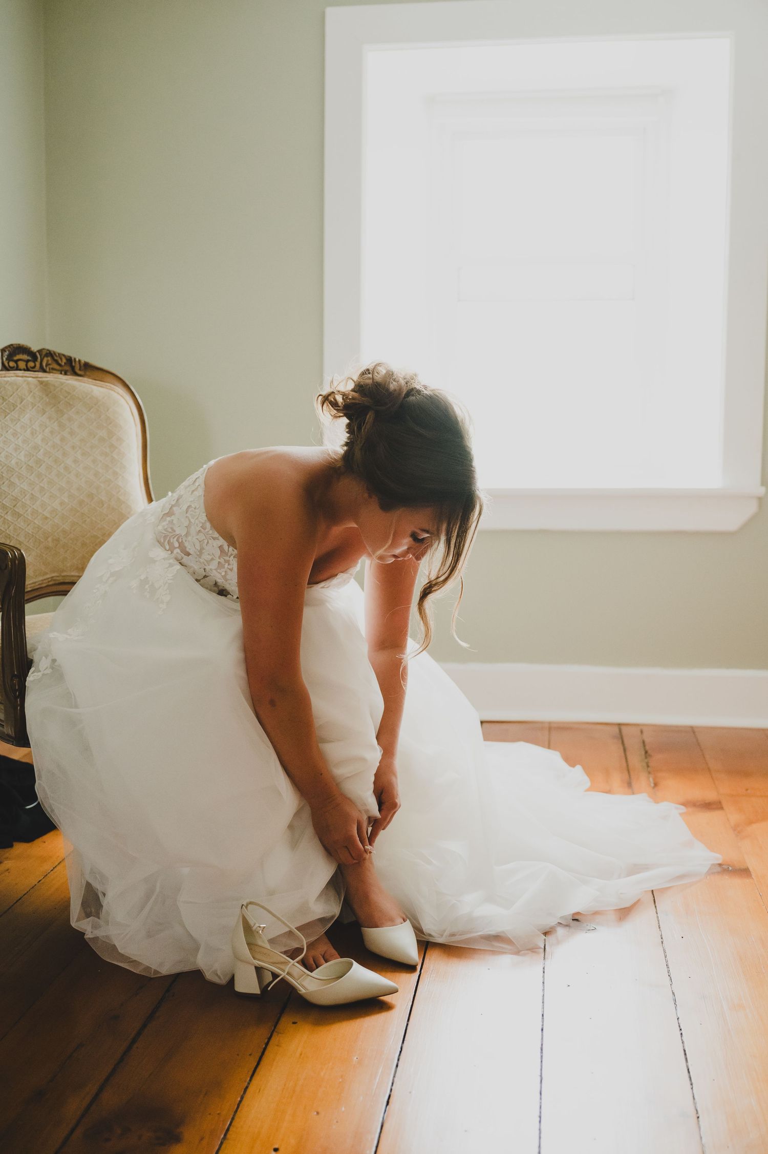 A person in a white wedding dress kneels down on wooden floorboards in a bright sunlit room.