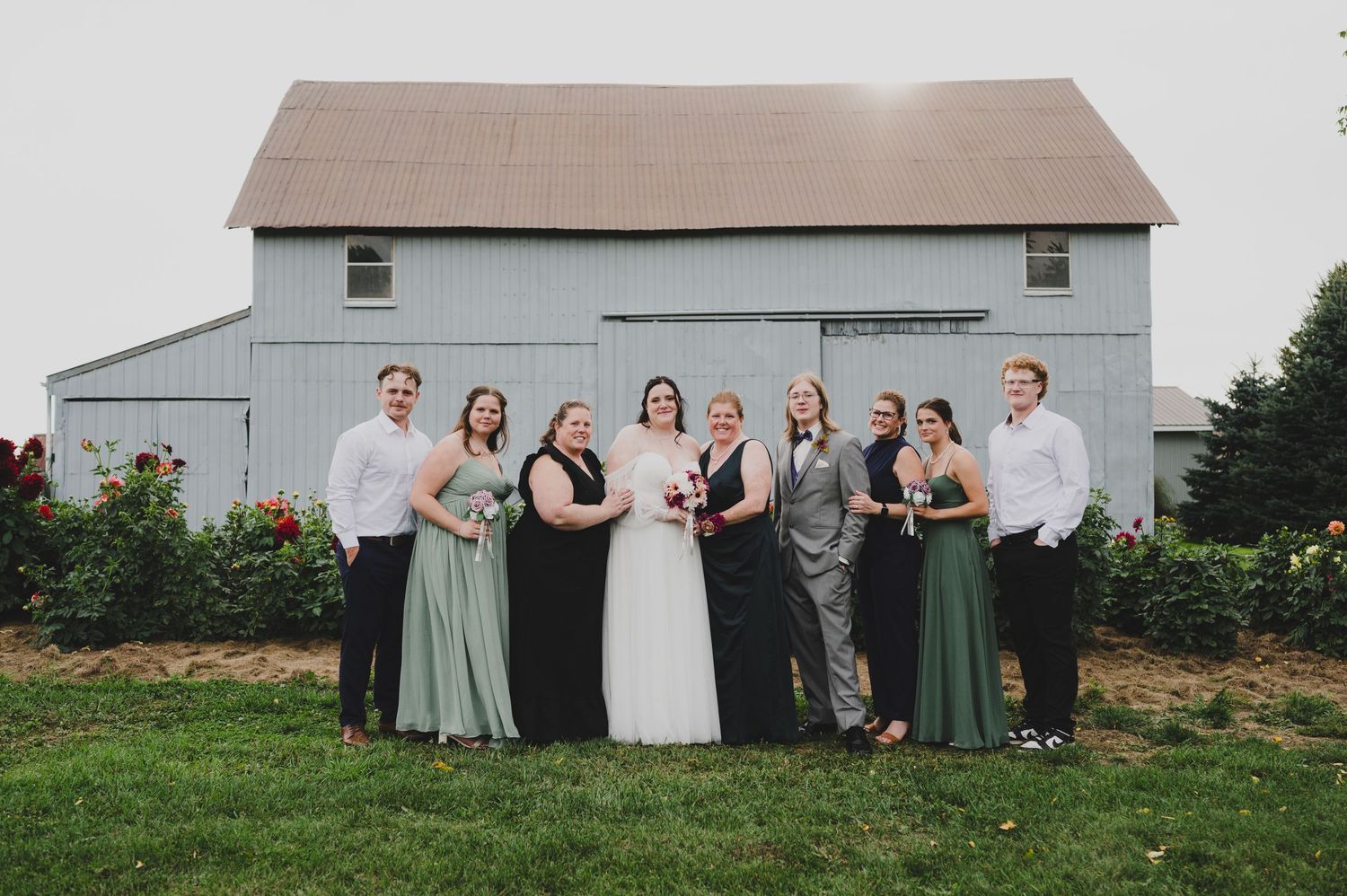 A wedding party poses for a group photo in front of a rustic white barn with flowers in the foreground.