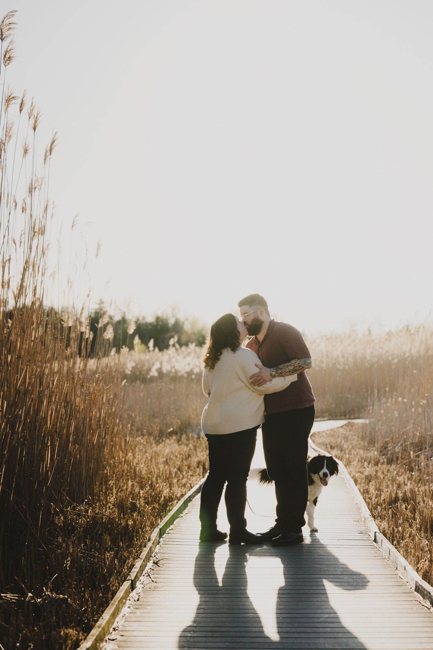 A couple stands together on a wooden boardwalk at sunset casting long shadows among tall reeds.
