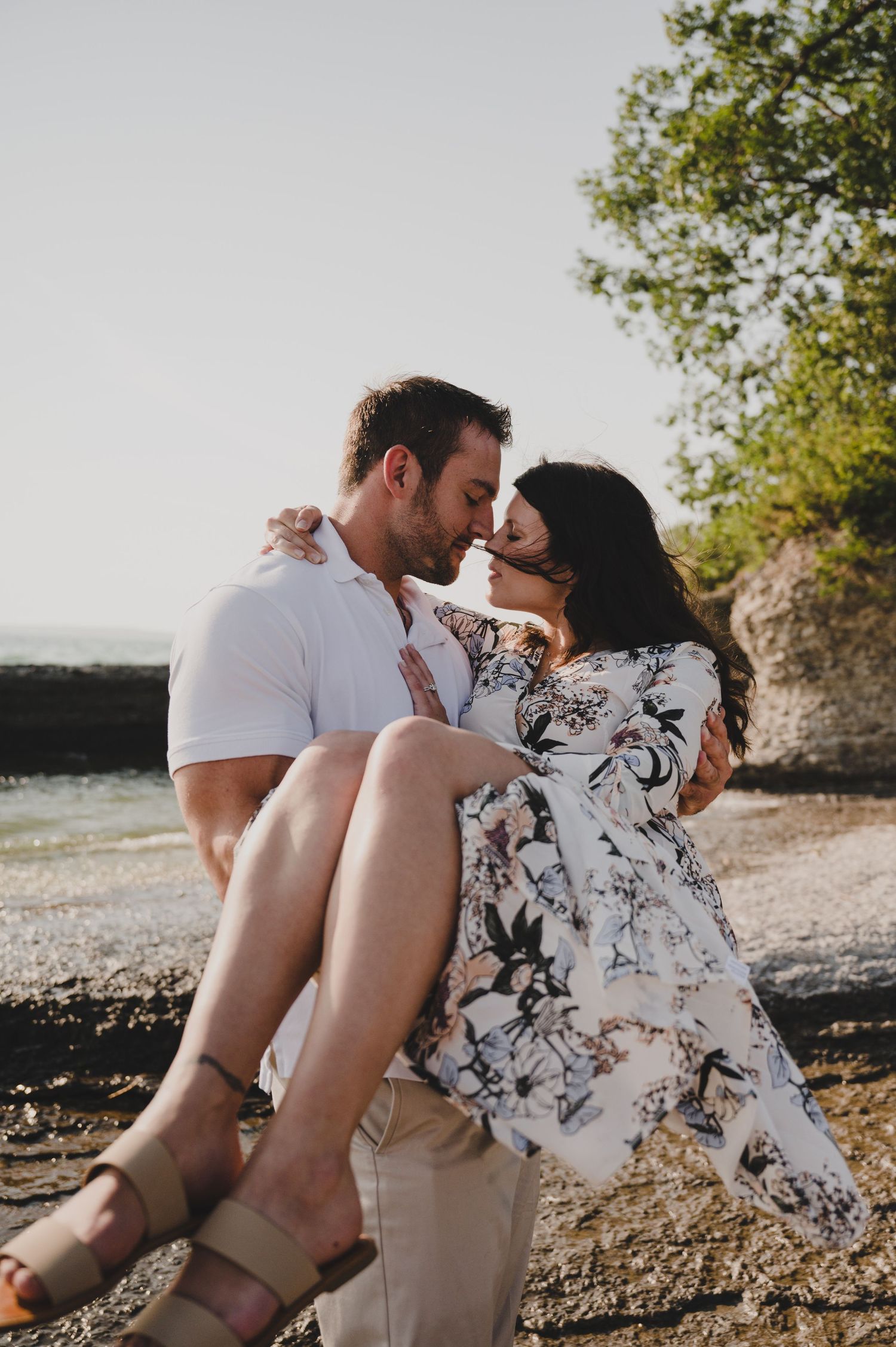 Romantic couple sitting together on a rocky beach at sunset sharing intimate moments in a floral dress and white shirt.