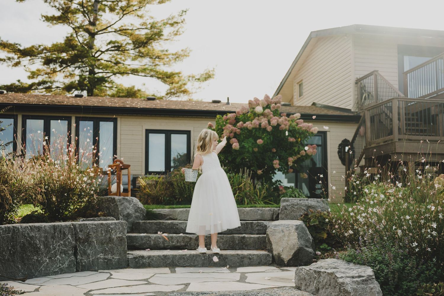 A white tulle dress stands elegantly on stone steps outside a modern house with lush garden landscaping.