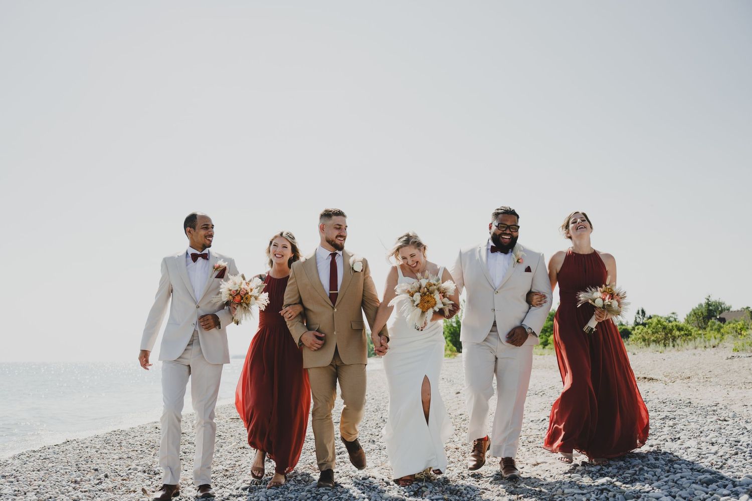 Wedding party walks on rocky beach, bride in white gown, groom in tan suit, bridesmaids in red dresses, groomsmen in white suits.