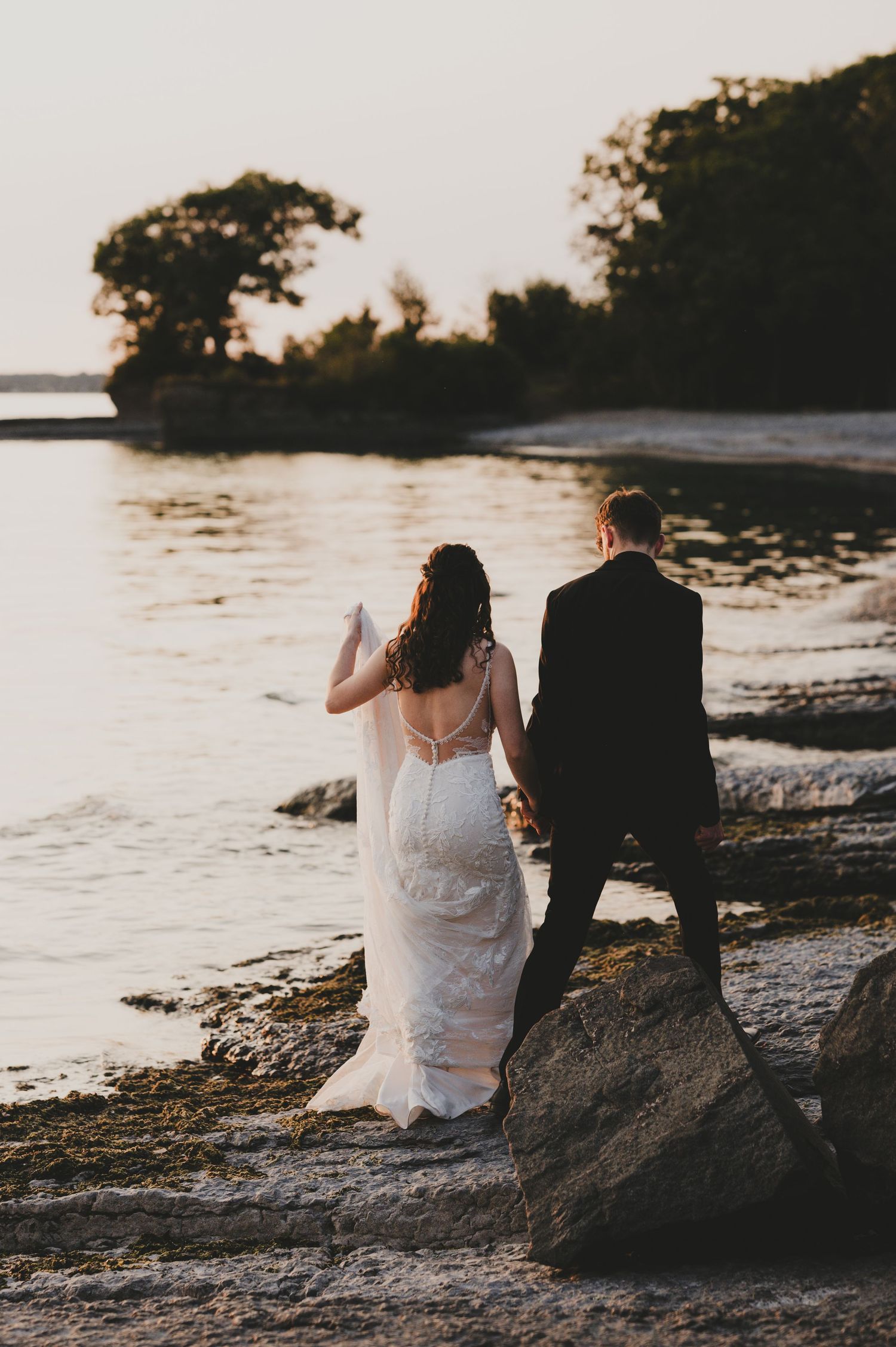Couple in wedding attire sits on rocky beach at sunset enjoying peaceful waterfront view.