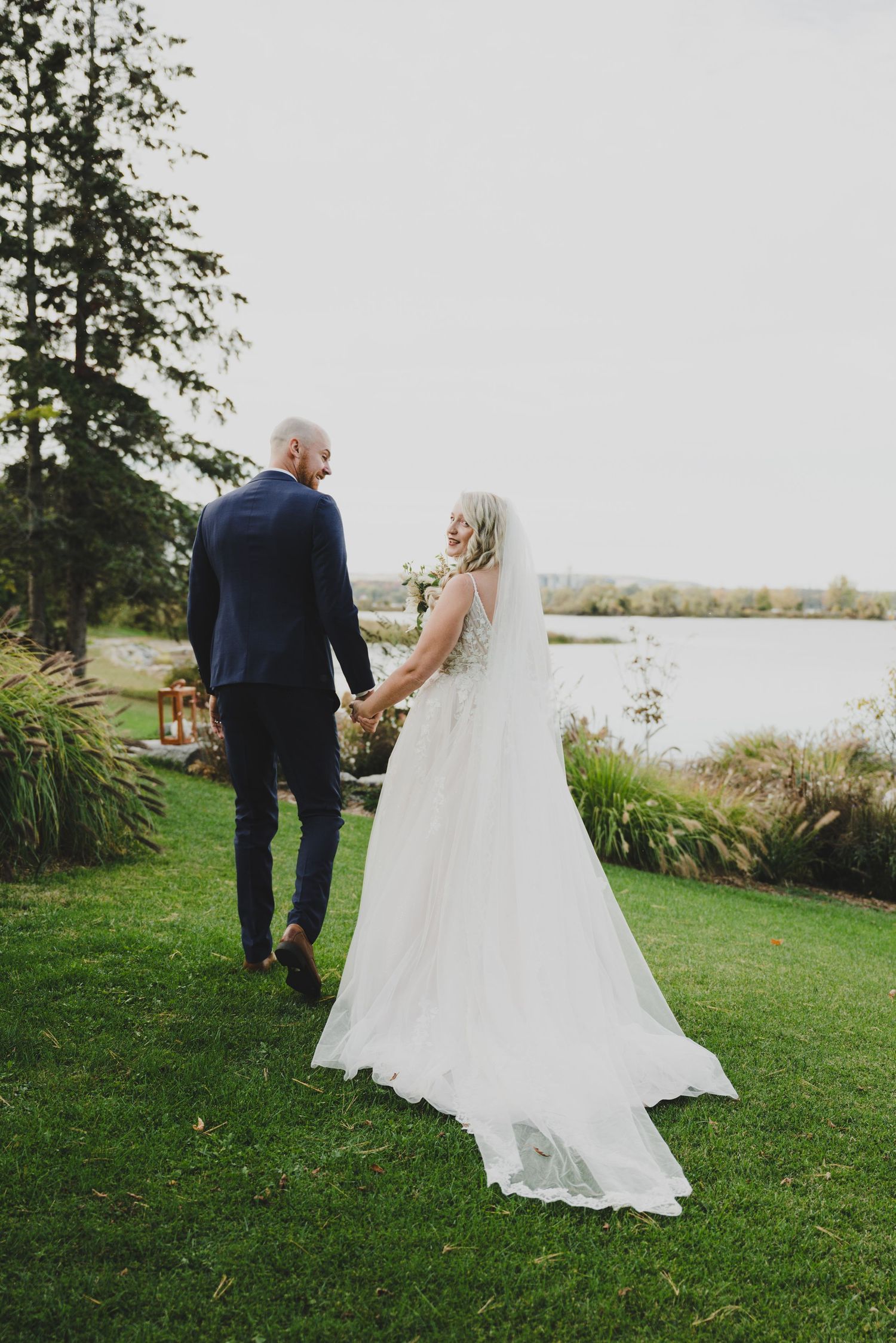 A couple in wedding attire holds hands while walking on grass near a serene lake during their outdoor ceremony.