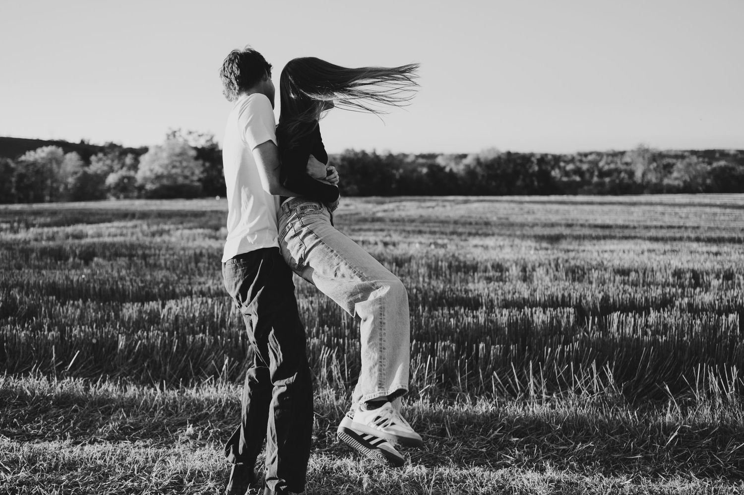 A couple in casual attire holds hands while walking through a scenic meadow in black and white.