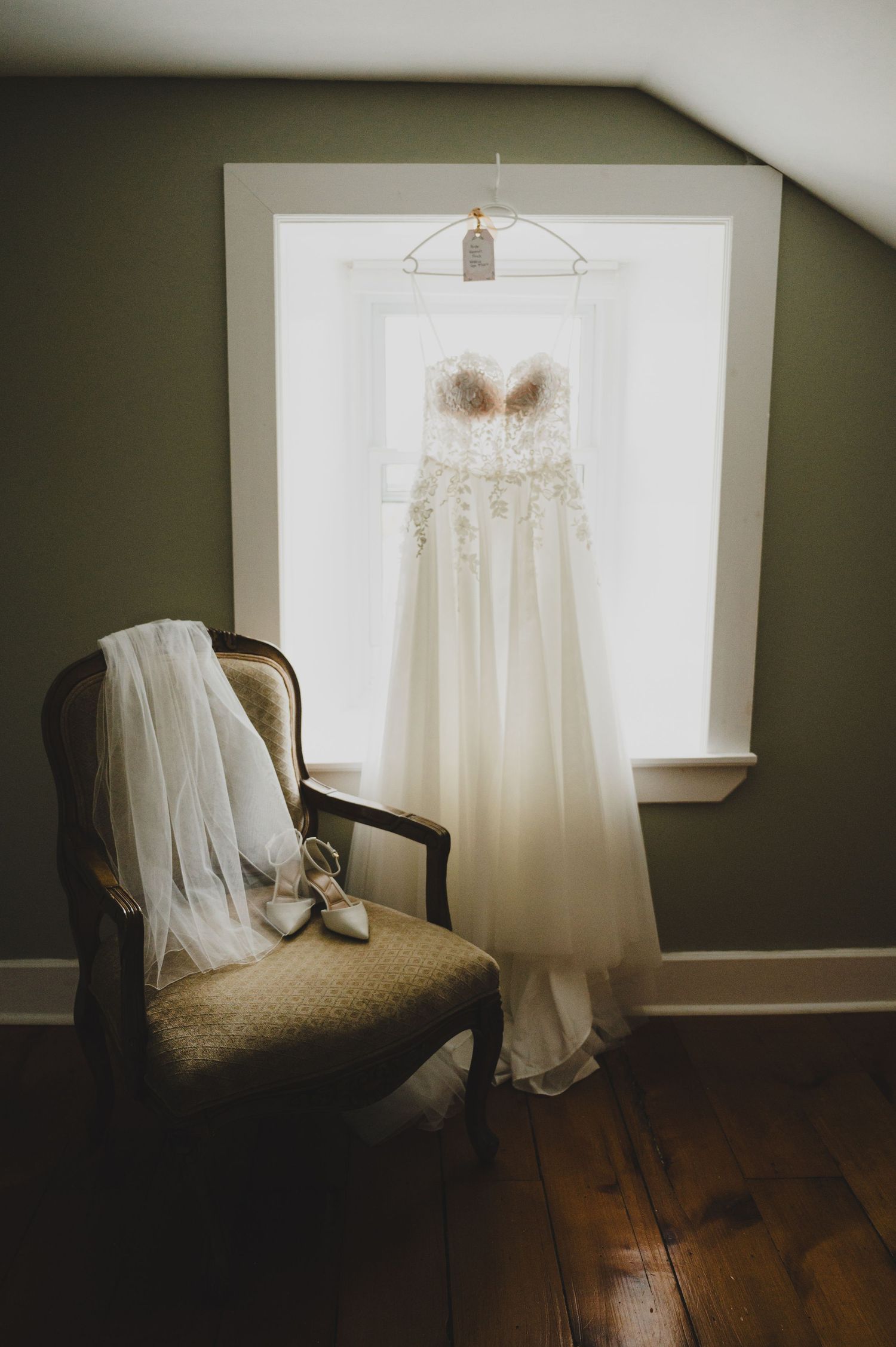 A white wedding dress hangs by a window with a veil and shoes draped over an antique chair on hardwood floors.