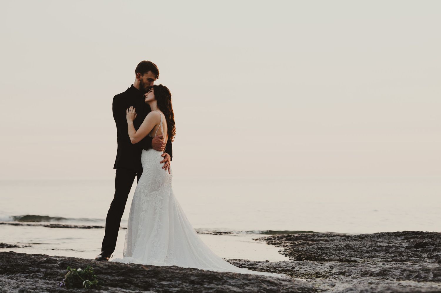 Wedding couple embraces on rocky coastal terrain during sunset.
