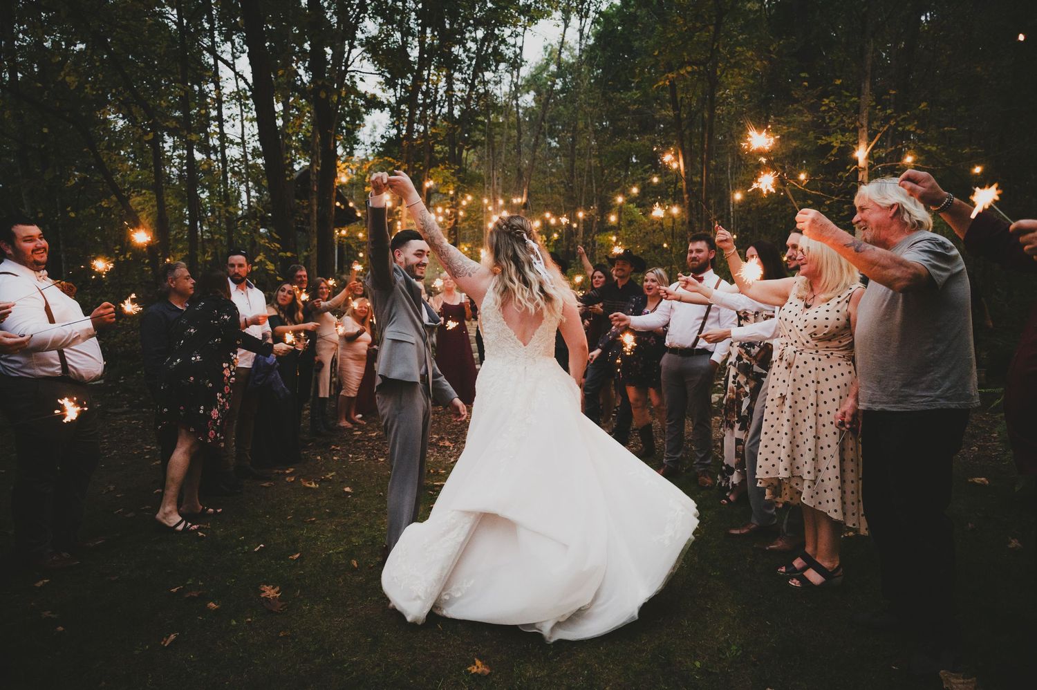 Wedding guests hold sparklers creating a magical archway during an outdoor evening celebration at dusk.
