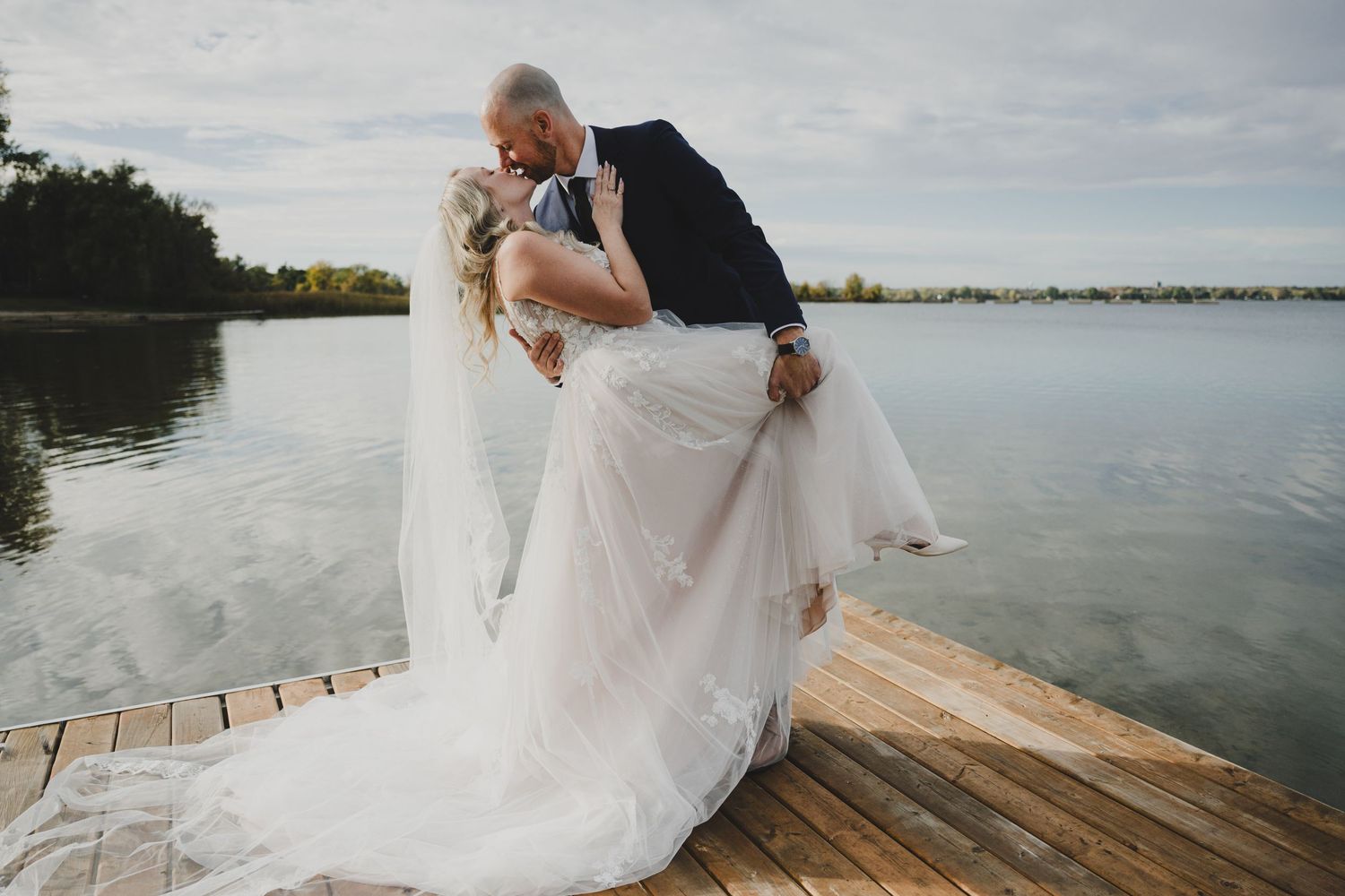 Bride and groom sharing a romantic kiss on a wooden dock by a serene lake, bride in white gown with veil.