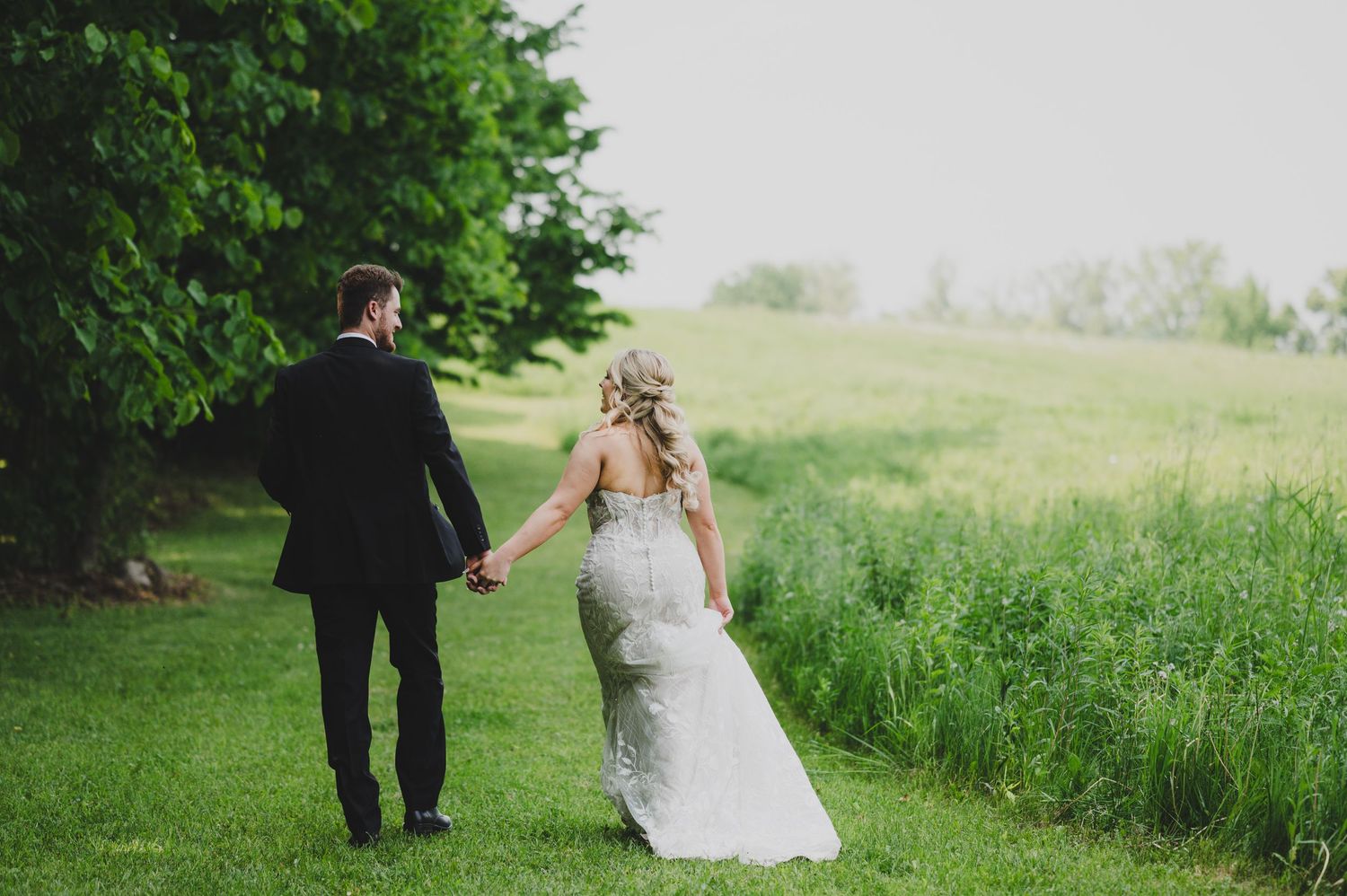 A couple in formal wedding attire walks through a lush green field on their special day.