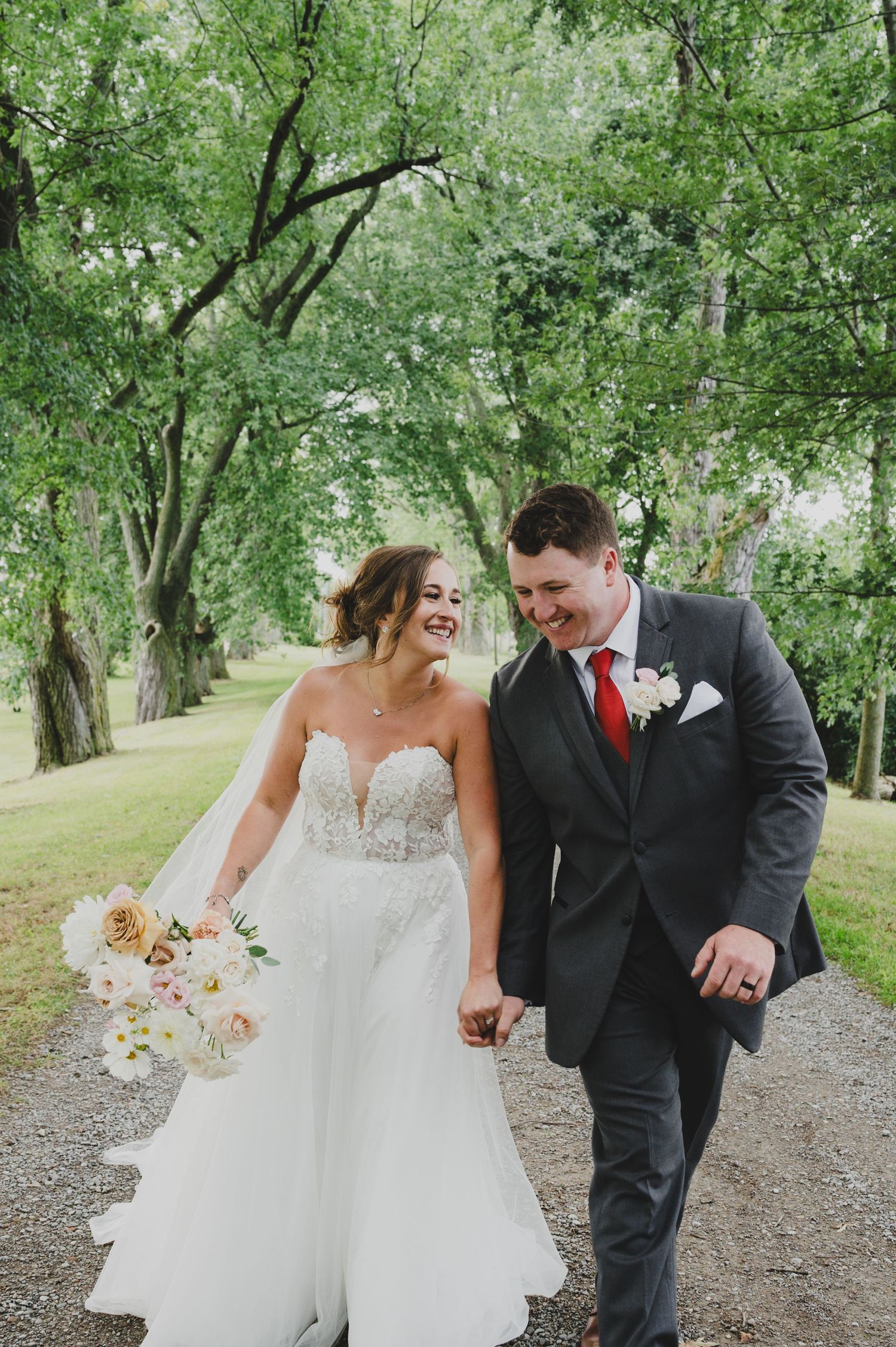 A couple walks hand in hand along a tree-lined path, the bride in white and groom in dark suit with red tie.