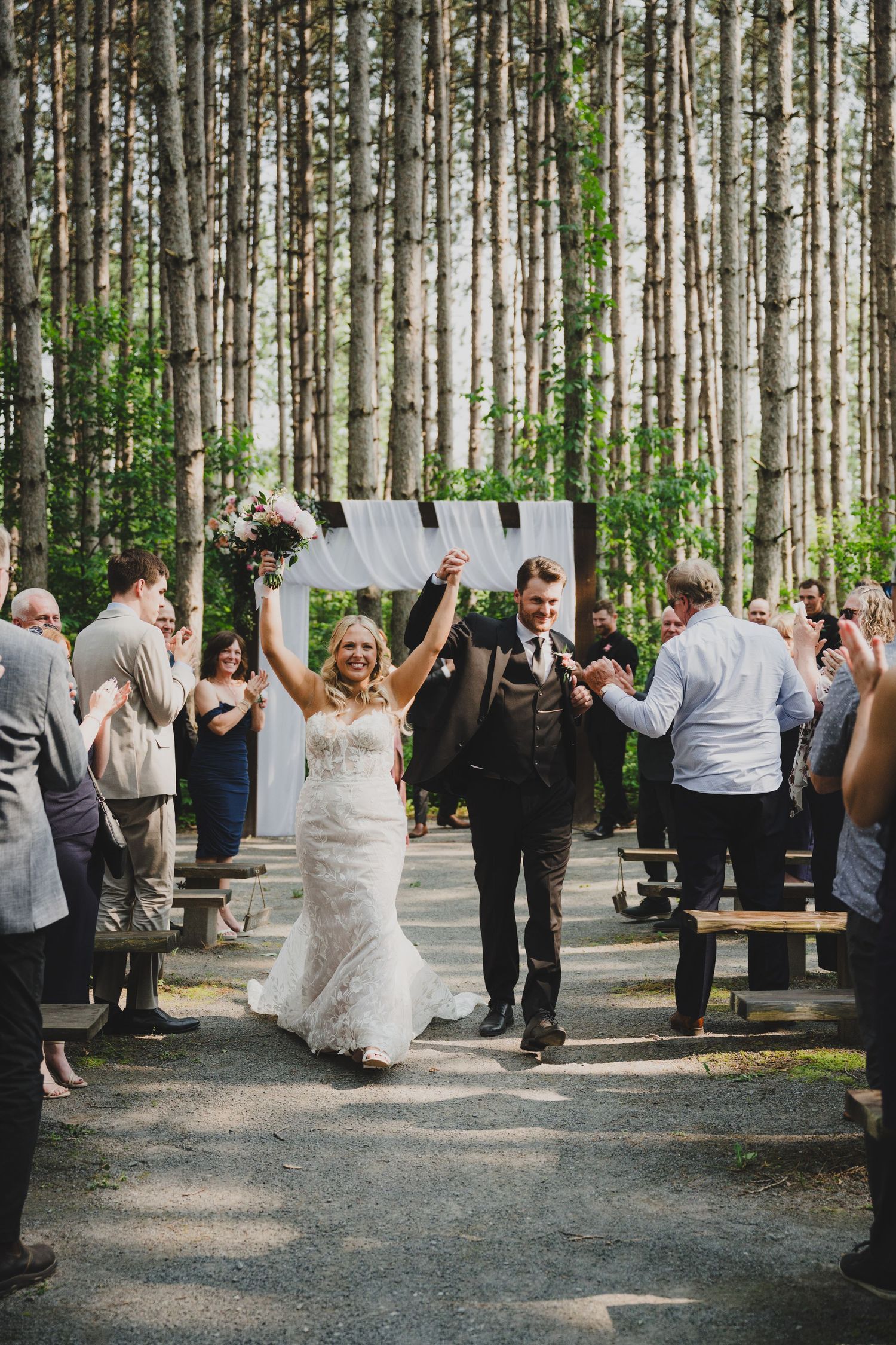 Wedding celebration in a pine forest with guests and couple dancing and mingling on a gravel path.