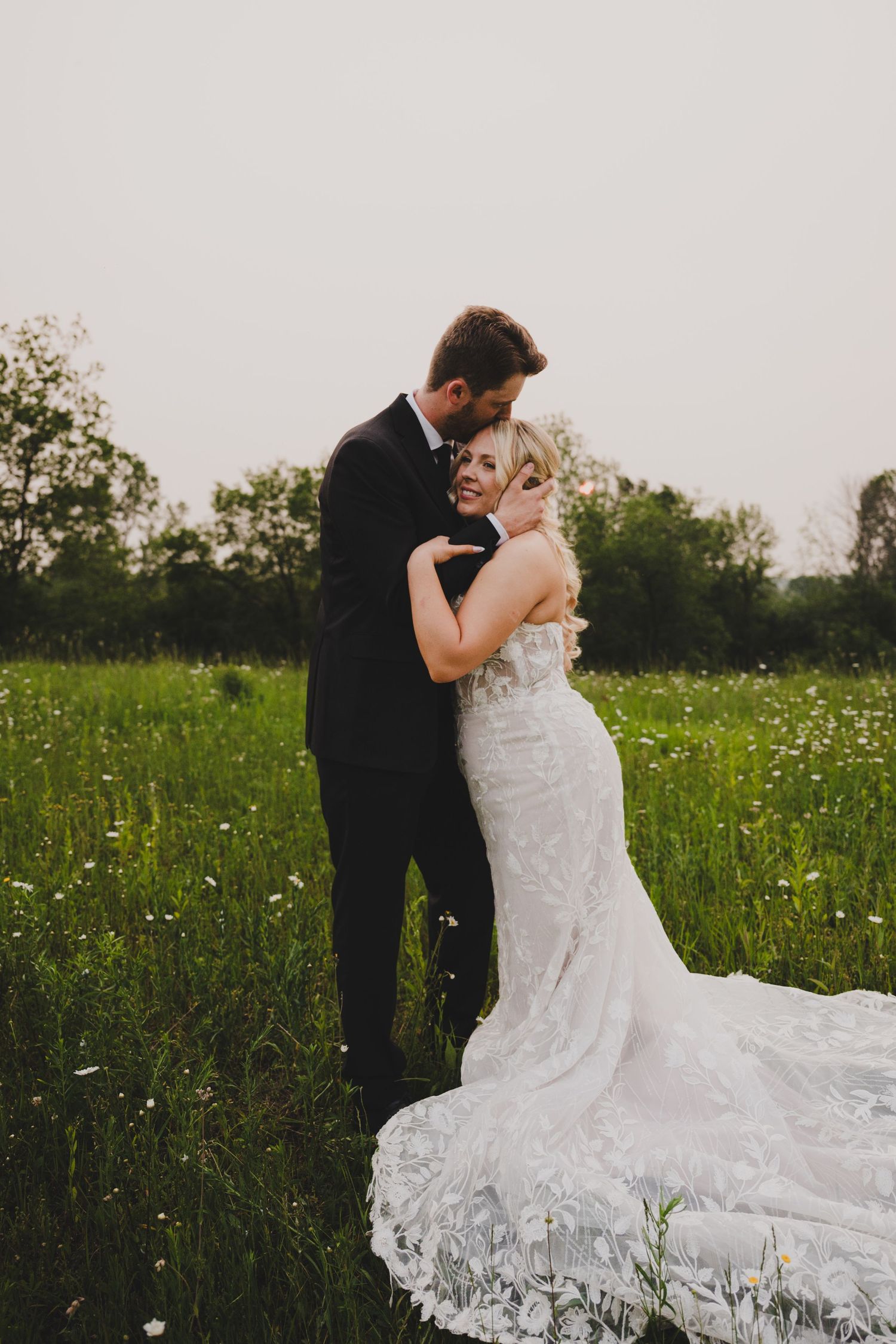 A romantic wedding couple dances in a wildflower meadow at sunset with the bride's lace dress flowing gracefully.