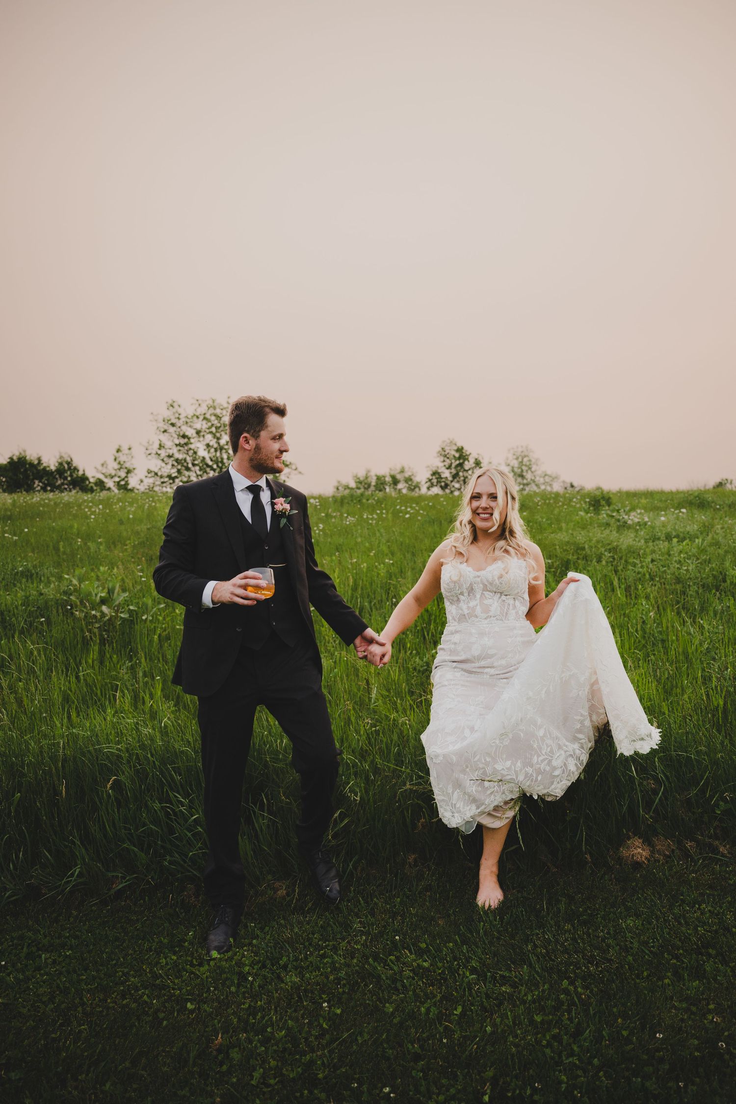 Newlywed couple sits playfully in a lush green meadow during their rustic outdoor wedding photoshoot.