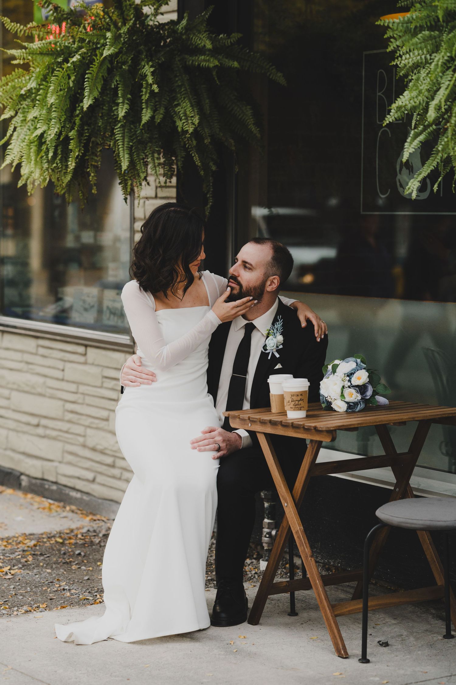 Couple sharing intimate moments on wooden bench outside cafe with lush greenery in background.