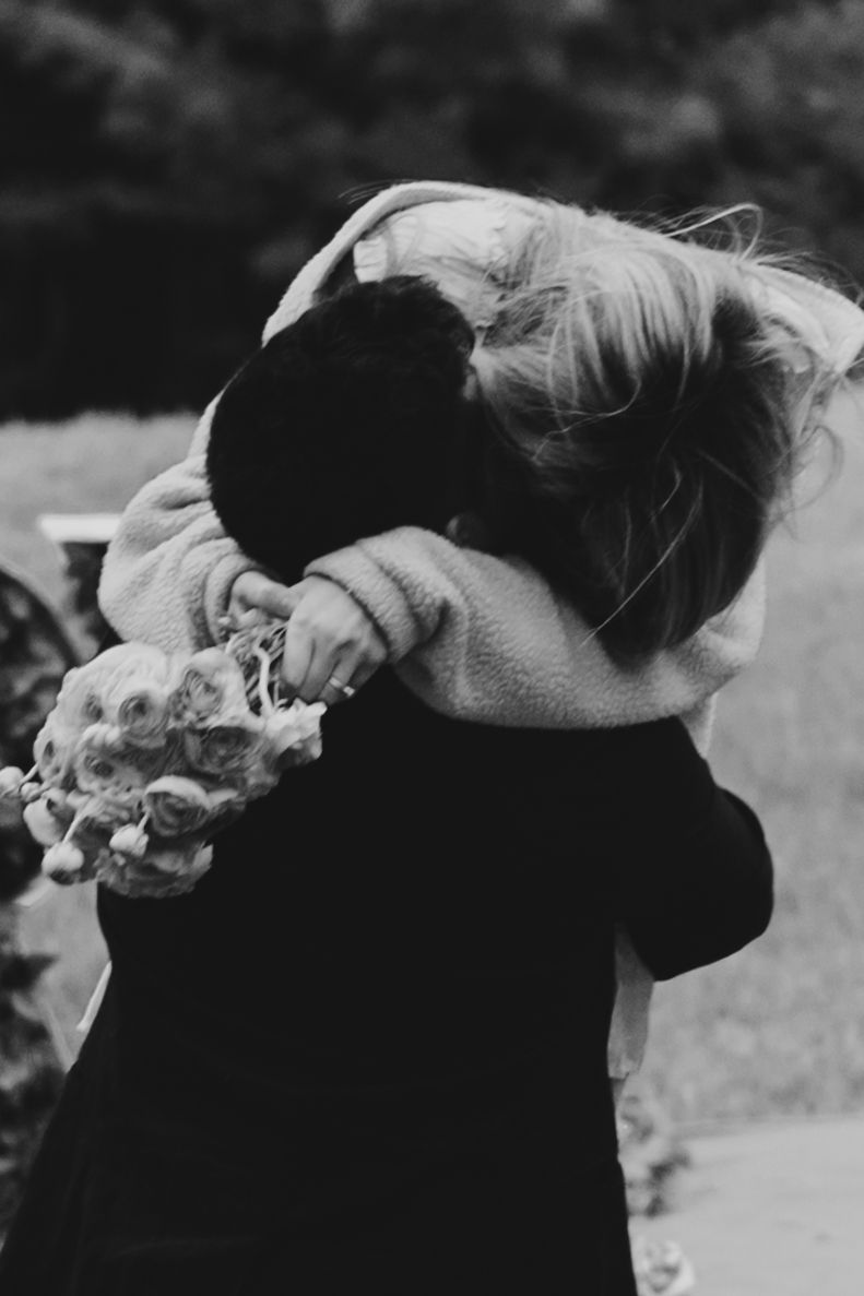 Two children share a tender hug outdoors, one holding flowers, captured in black and white photography.