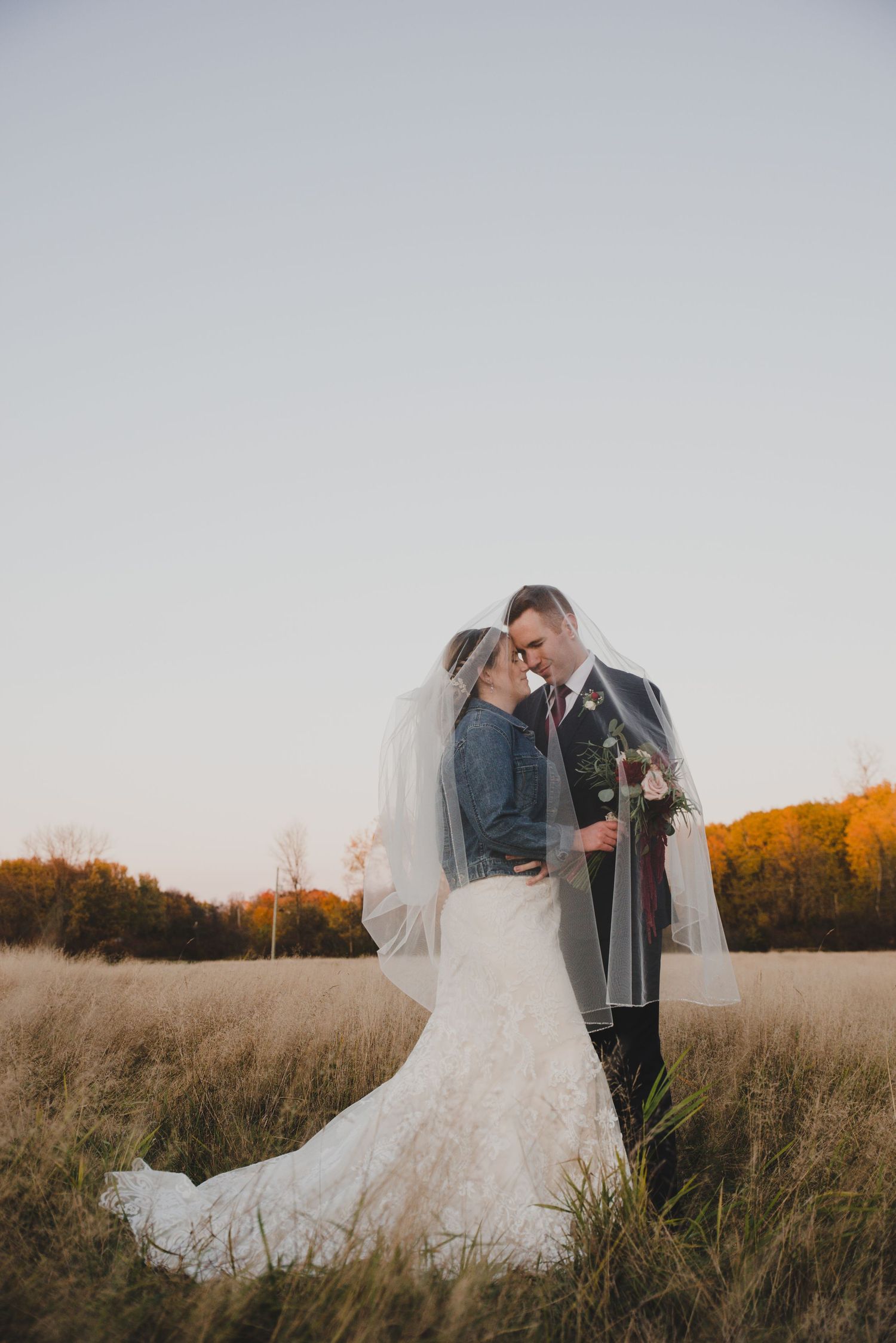 Bride and groom share romantic moment under veil in golden autumn field at sunset, bride in lace gown with denim jacket.