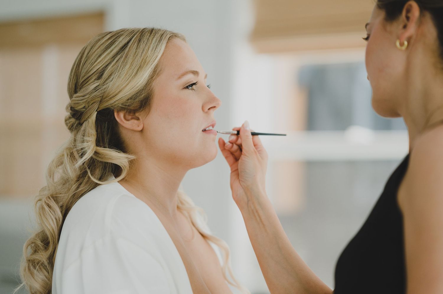 Professional makeup artist applies lipstick to a bride during wedding day preparations in natural light.