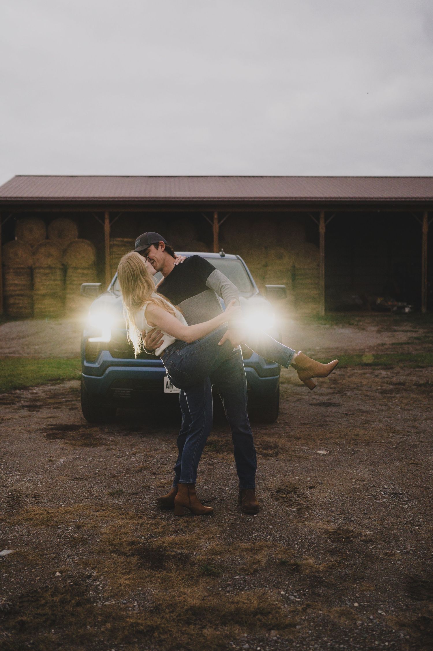 A romantic couple embraces in front of a pickup truck with glowing headlights at dusk near a rustic barn.