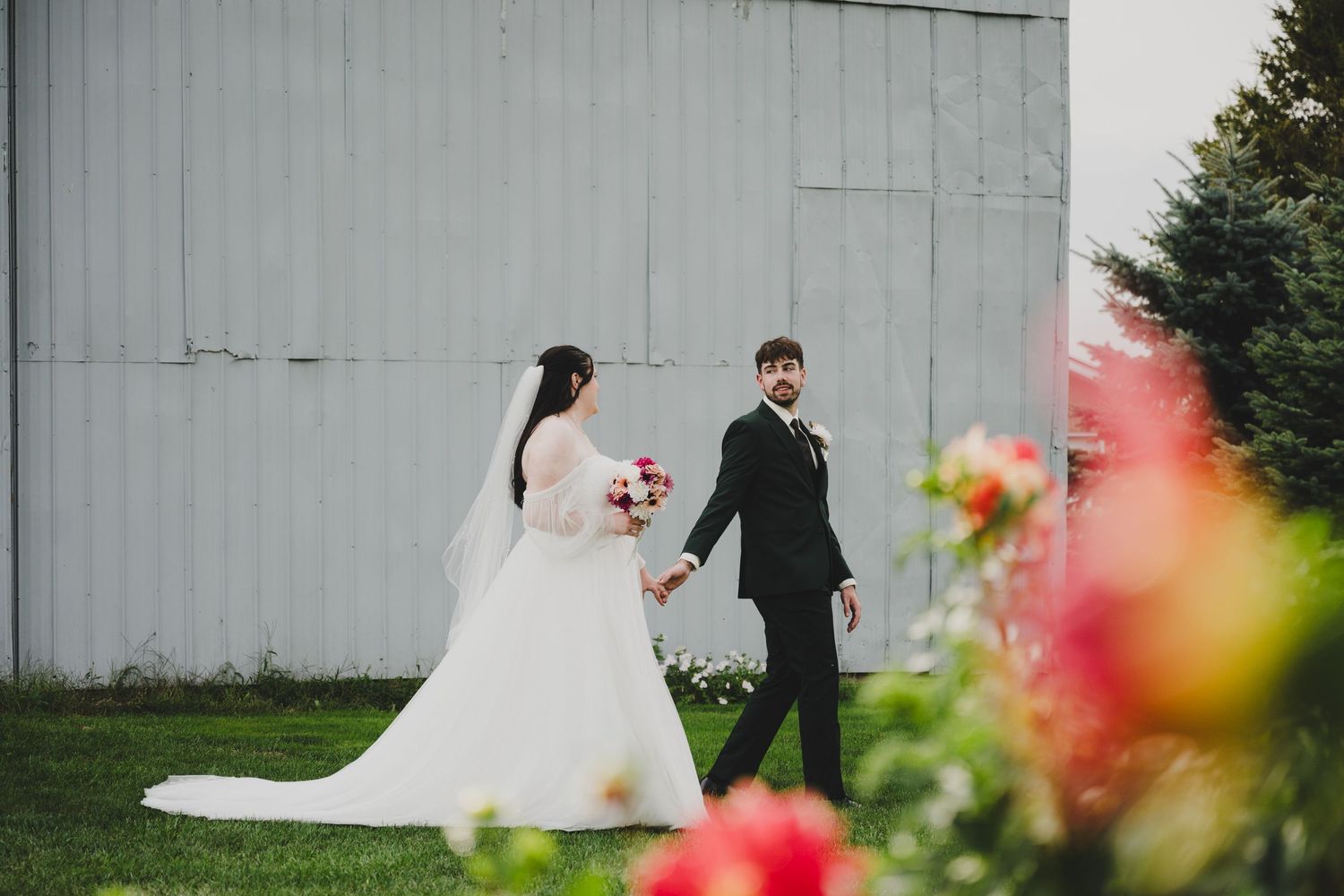 A couple in wedding attire walks along a rustic white barn wall with colorful flowers in the foreground.