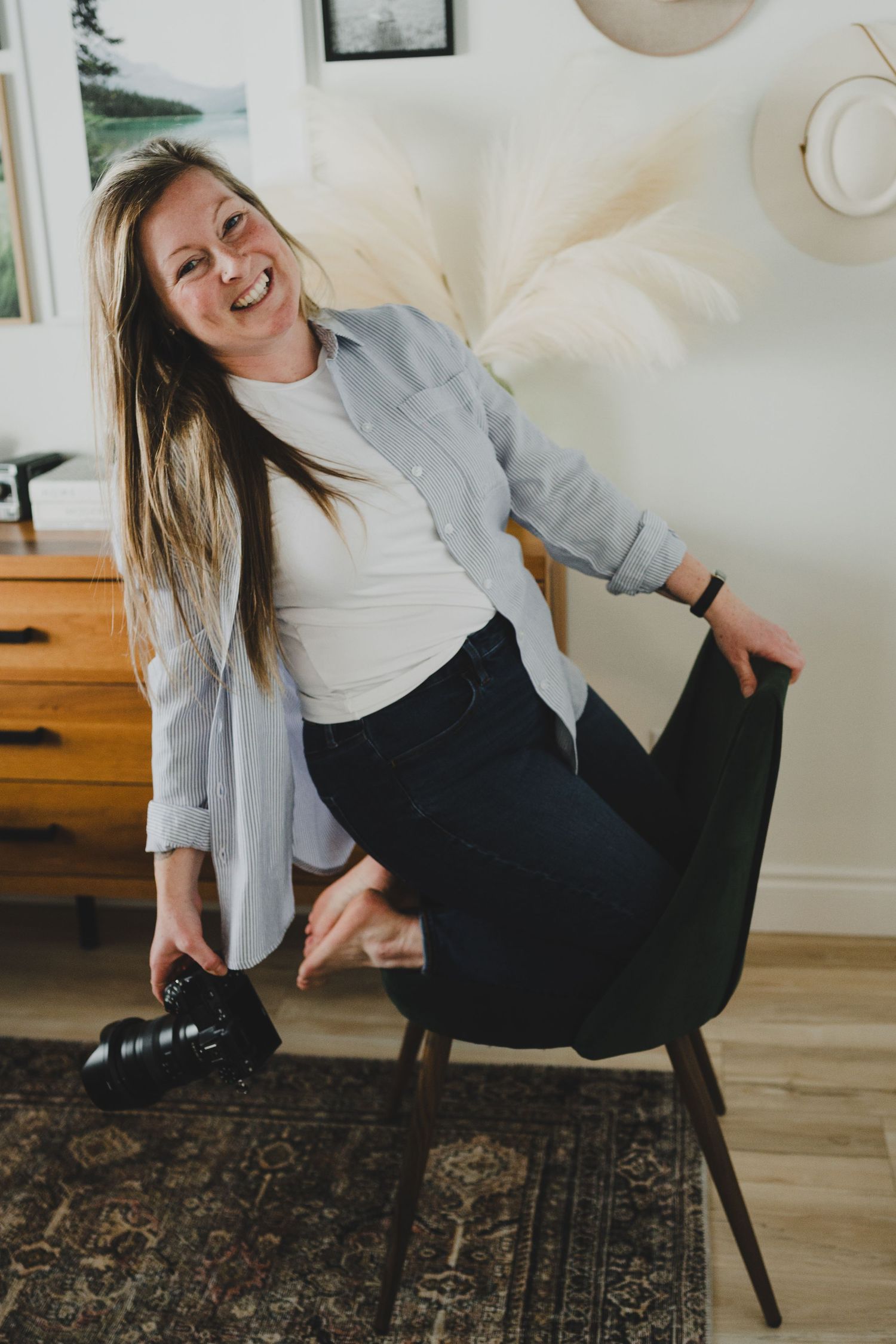 Smiling woman in gray shirt and dark jeans sitting on a modern chair holding a camera in a cozy home interior.