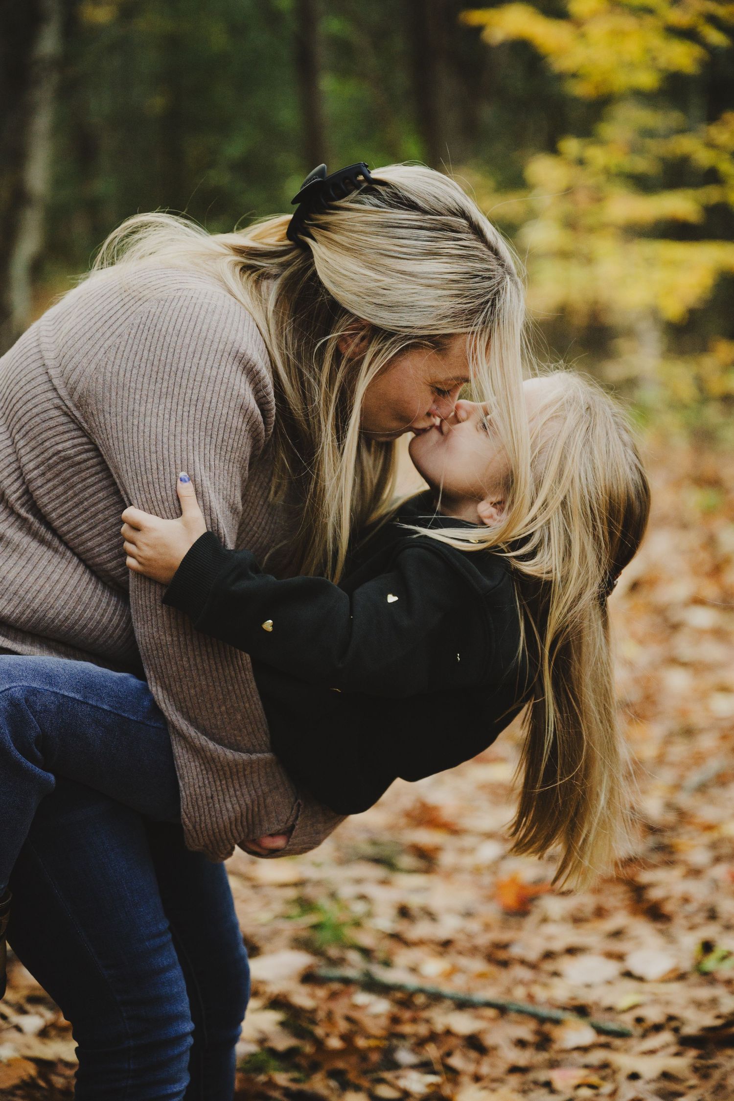 Person in brown sweater and jeans bends down in autumn forest with fallen yellow leaves.