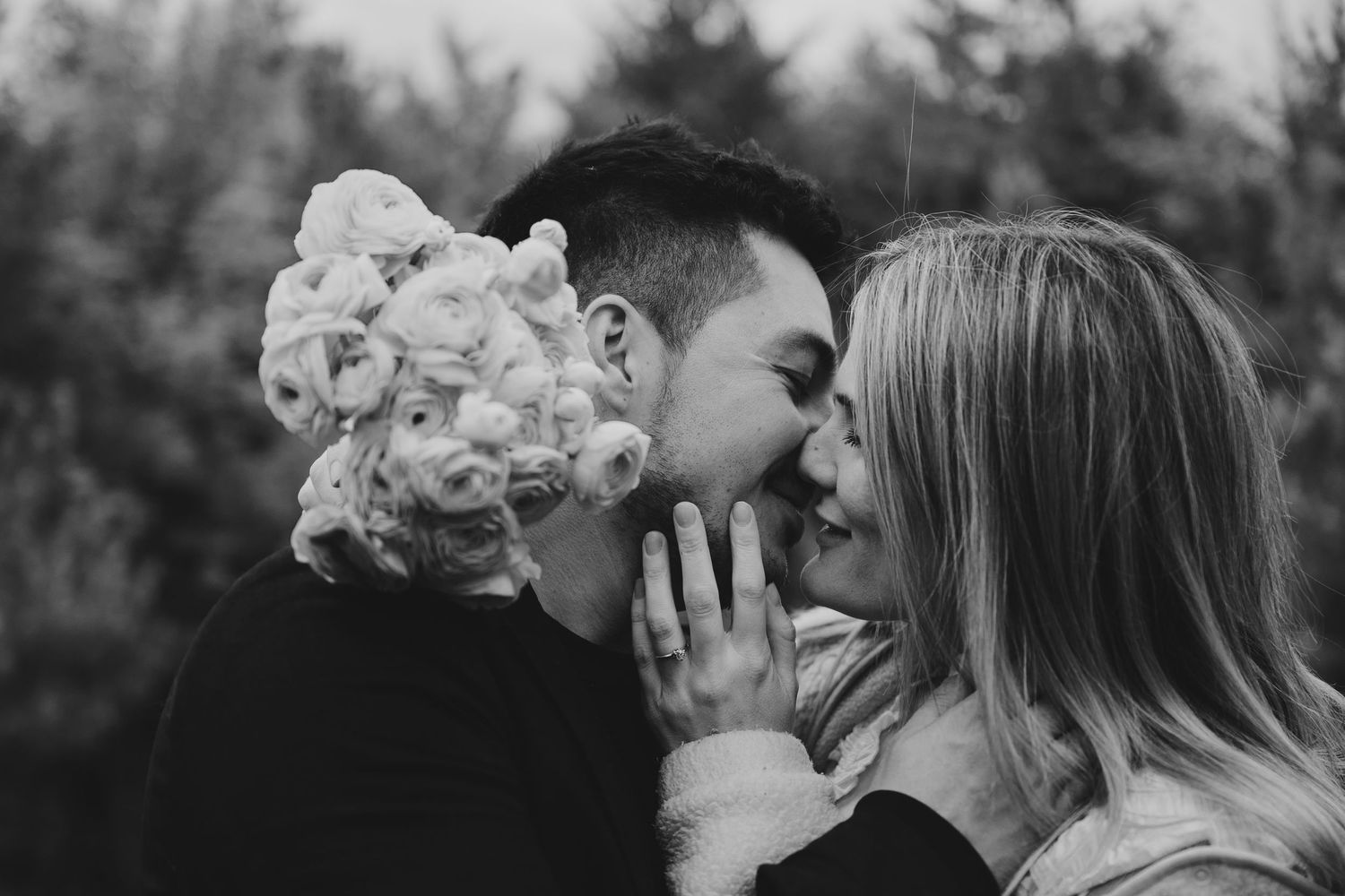 Black and white photo of two people sharing a kiss outdoors, with a person wearing a floral headpiece nearby.