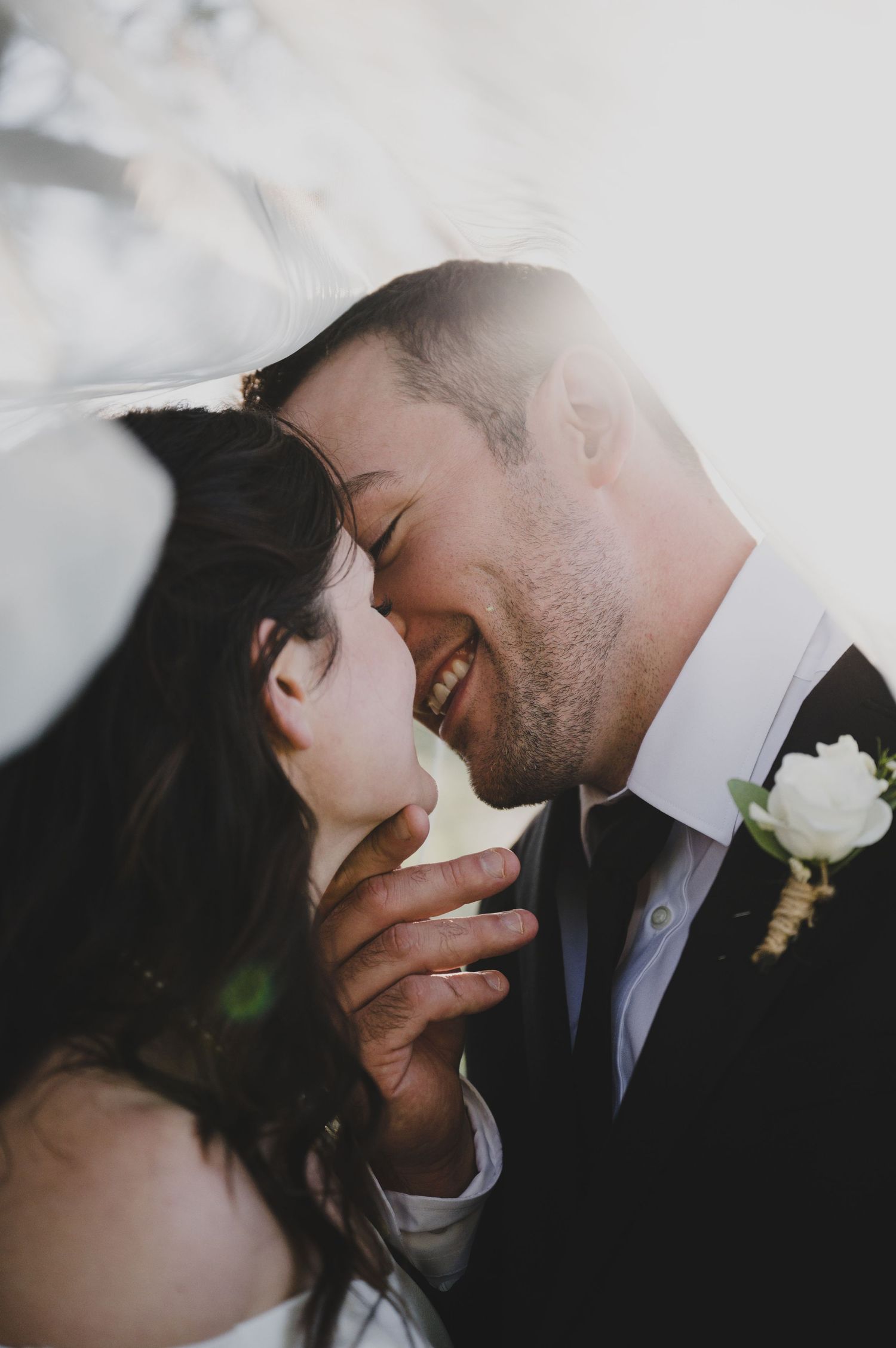 Intimate moment between newlyweds with white boutonniere and soft sunlight.