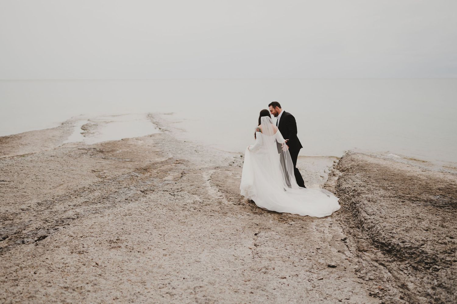 Bride and groom embrace on rocky shoreline with misty horizon, bride's long veil trailing behind in elegant wedding portrait.