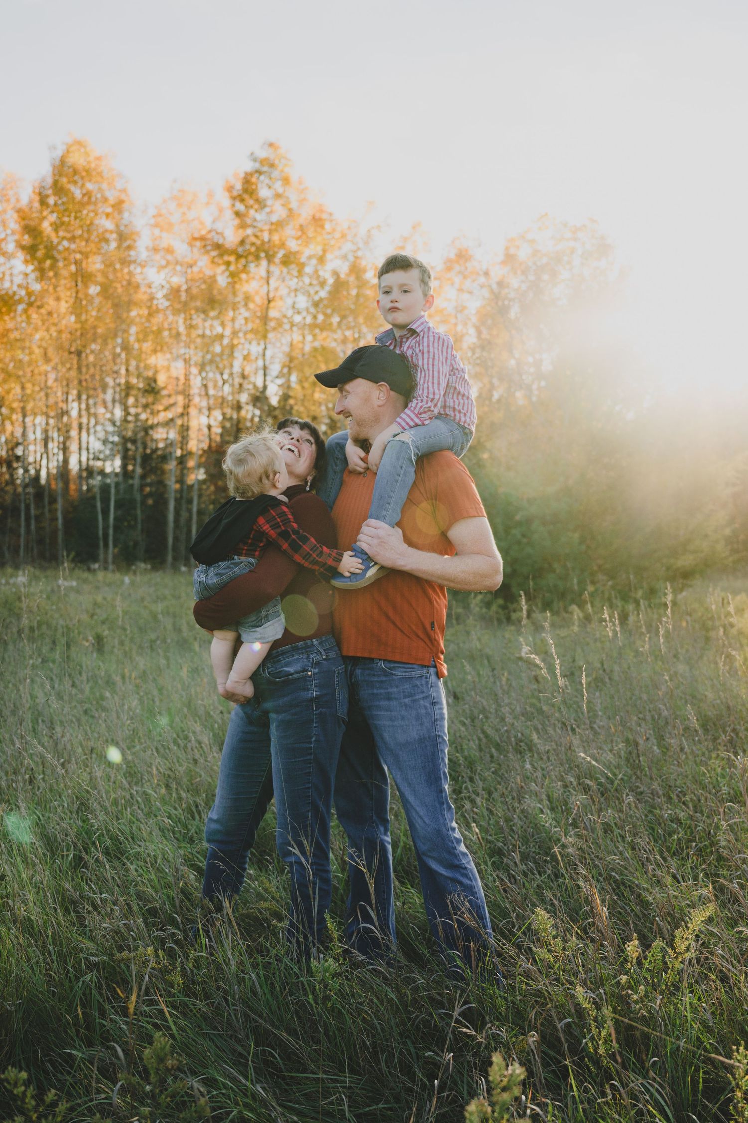Family enjoying tender moments together in a sunlit meadow during golden hour with autumn trees in background.