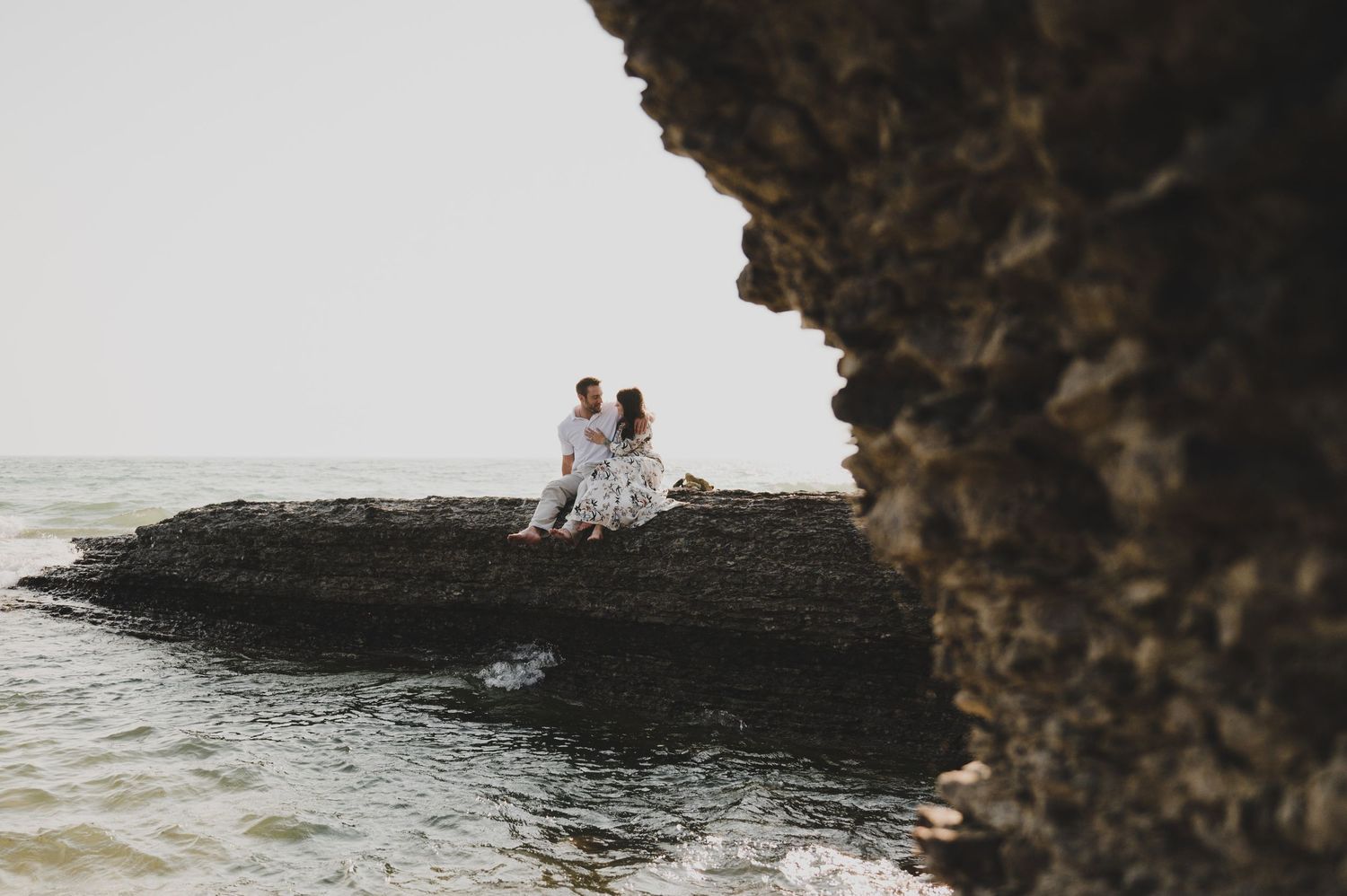 Two people sit together on a rocky ocean cliff overlooking the water at sunset.