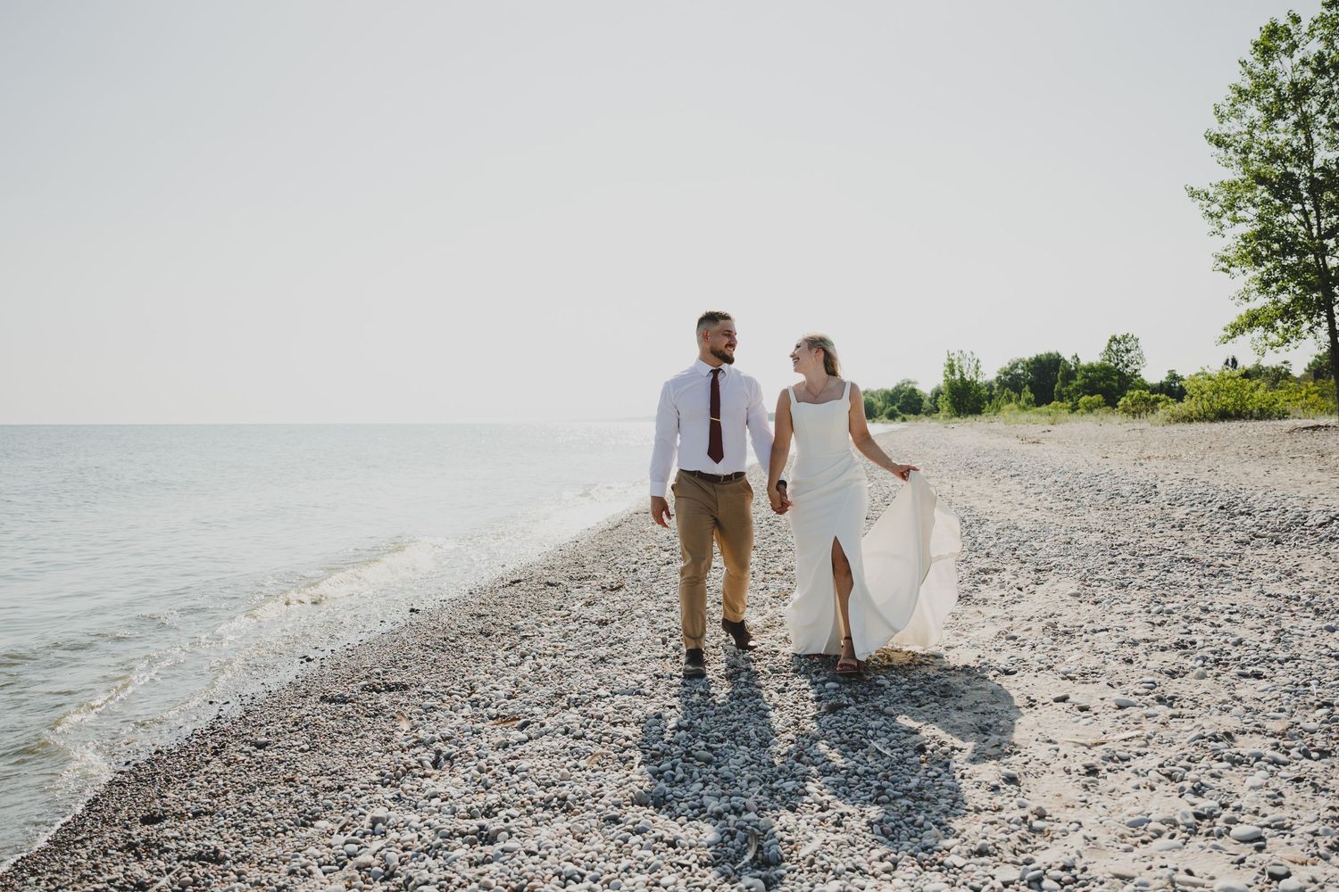 A couple walks hand in hand along a rocky beach shoreline on a sunny summer afternoon.