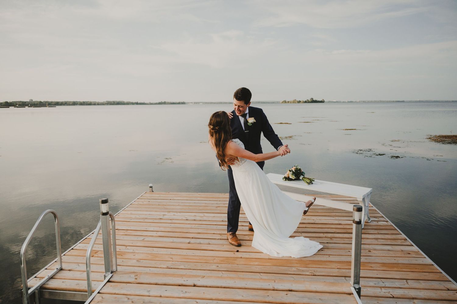 Bride and groom dancing on a wooden dock overlooking a calm lake on their wedding day.