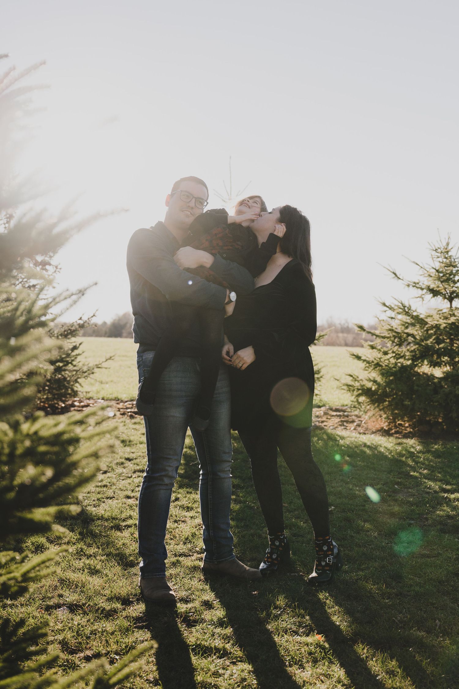 A family embraces together outdoors on a sunny day with evergreen trees and lens flares in the background.