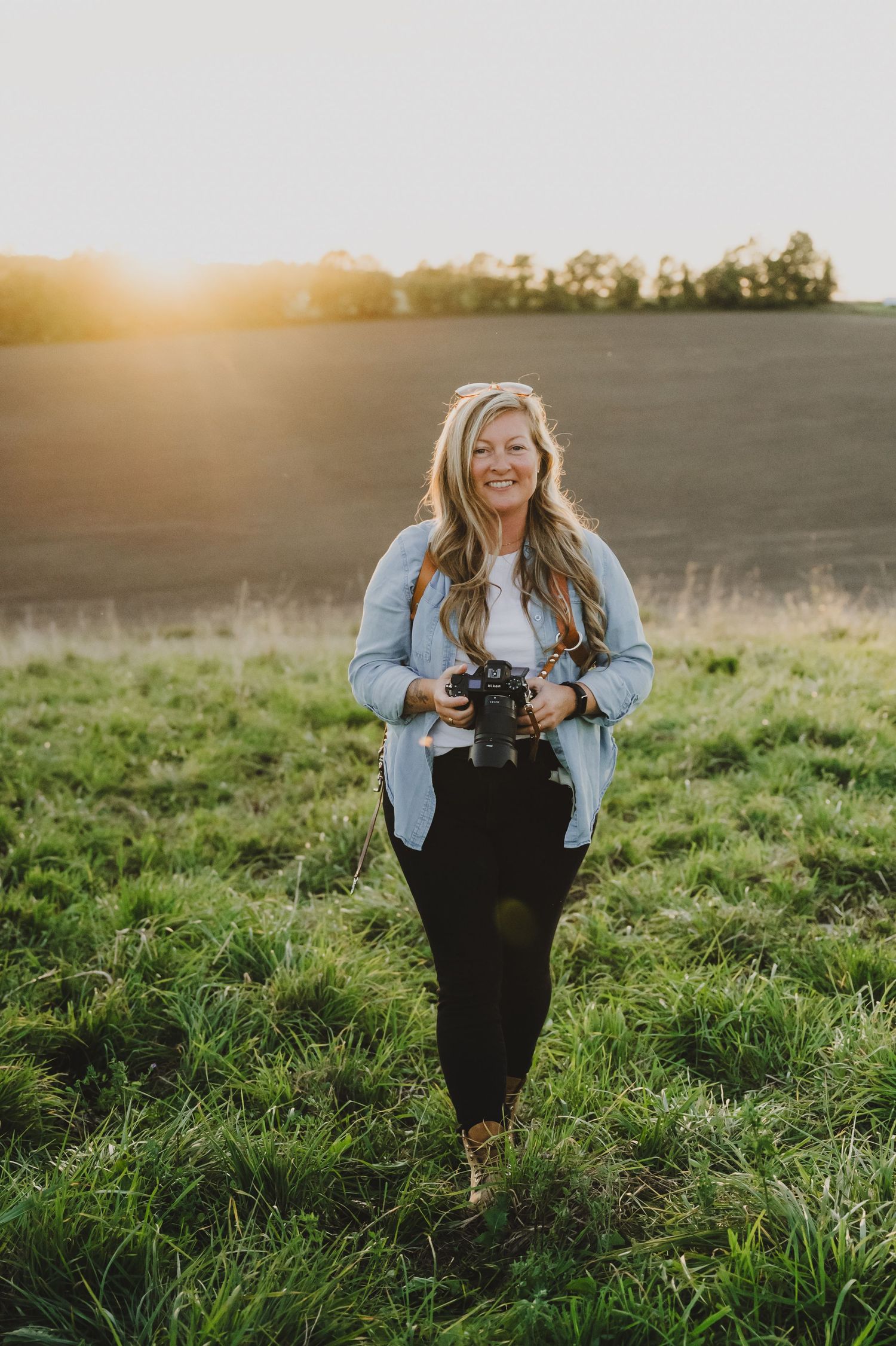 A photographer in a denim jacket and dark dress sits in a grassy field during golden hour sunset.