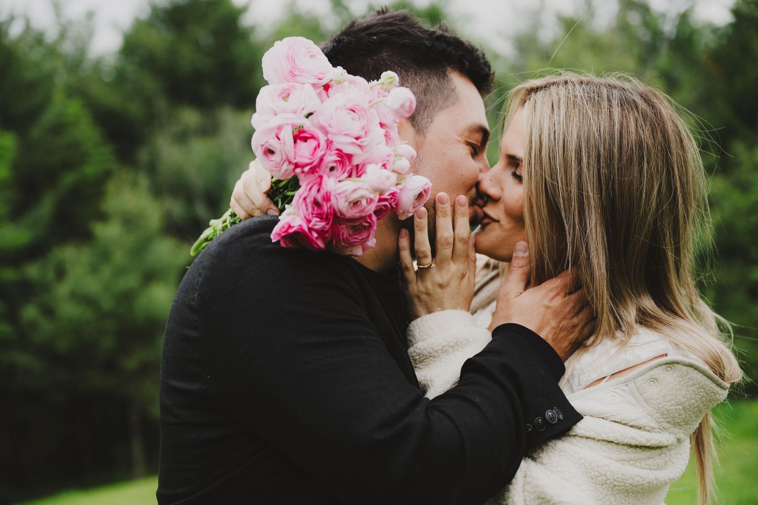 A couple shares a romantic kiss in a lush garden setting with pink peonies and greenery in the background.