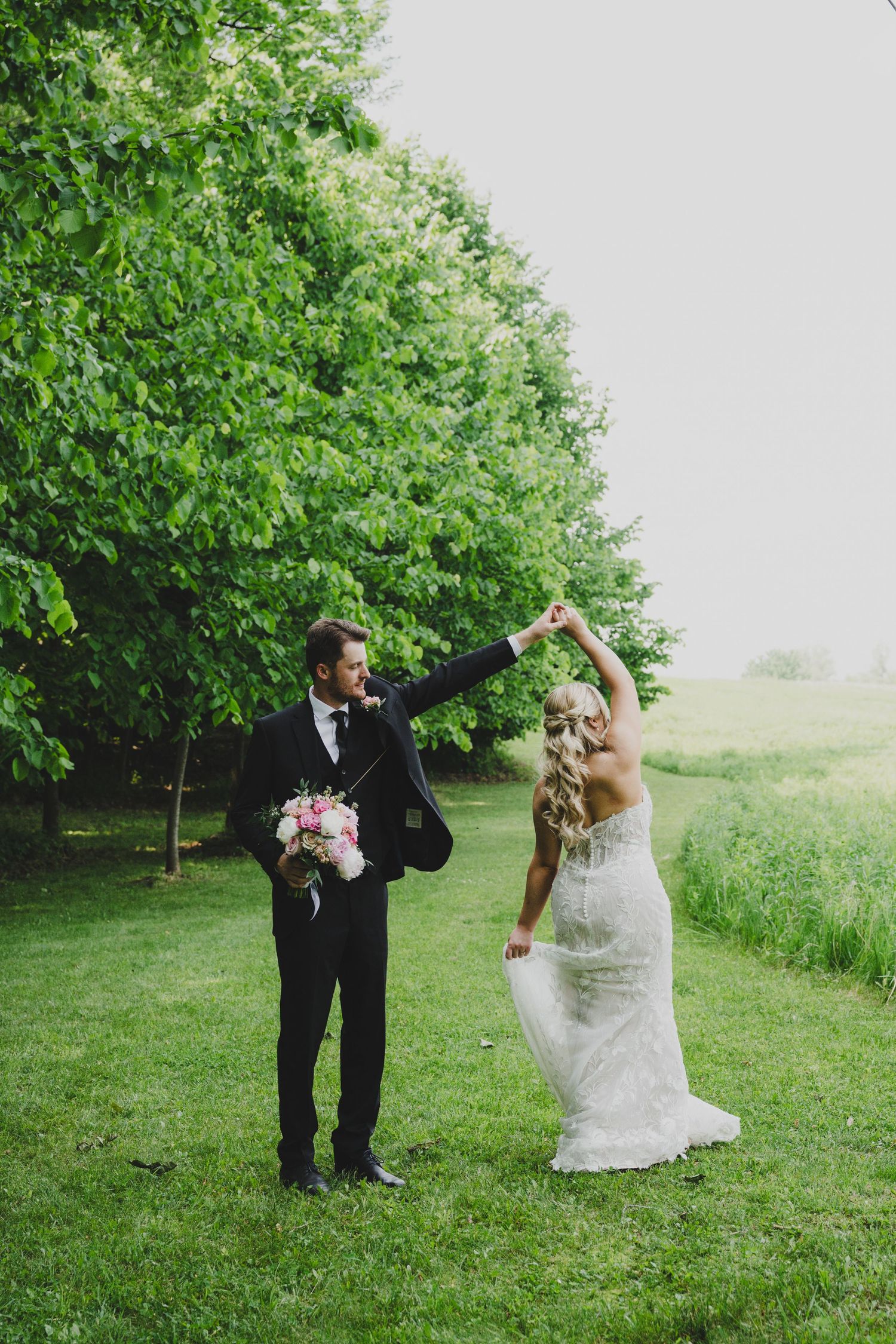 Couple in wedding attire dancing and twirling together on a lush green lawn surrounded by trees.