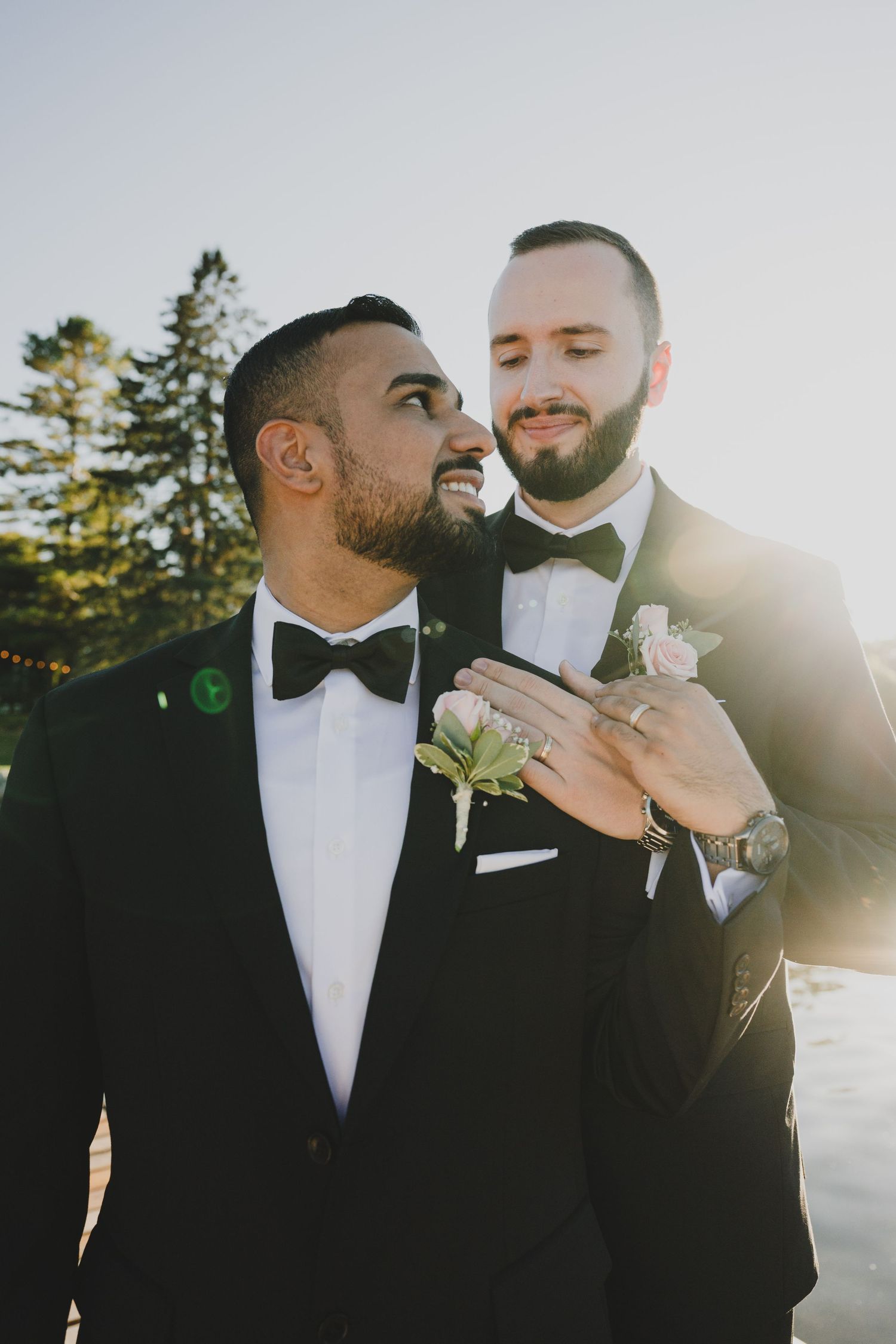 Two people in black tuxedos and bow ties share an intimate moment in golden sunlight during a wedding celebration.