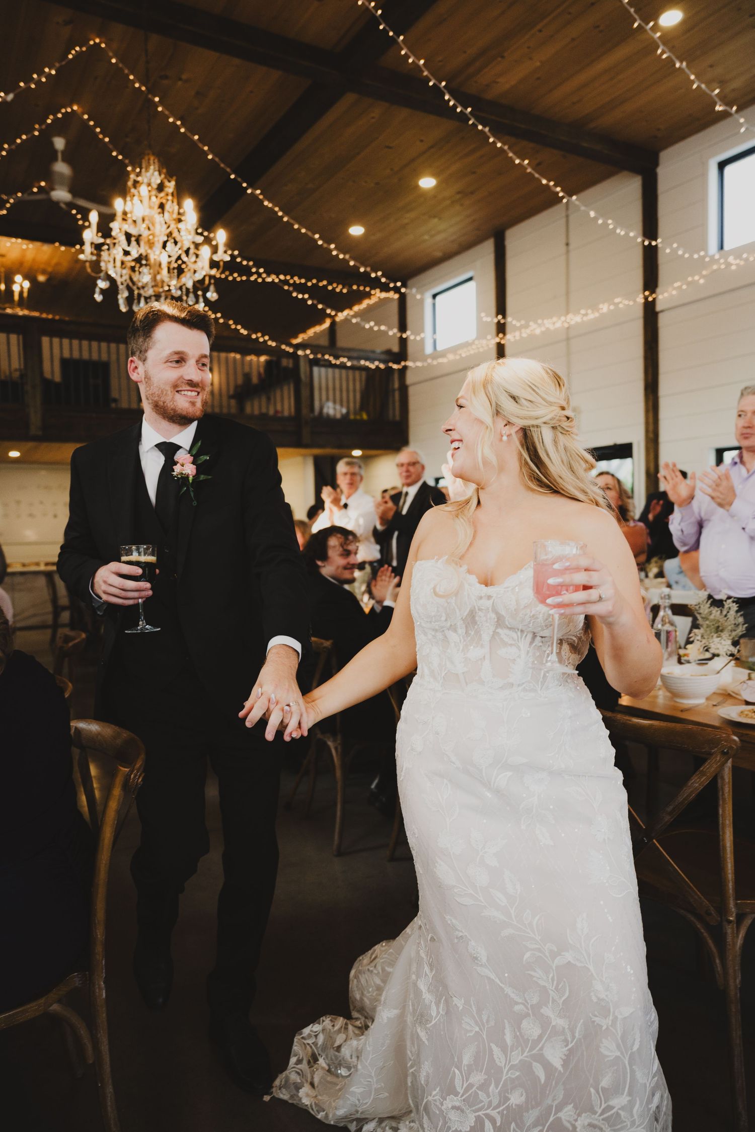 Wedding reception guests enjoy drinks and conversation under twinkling string lights in a rustic venue.