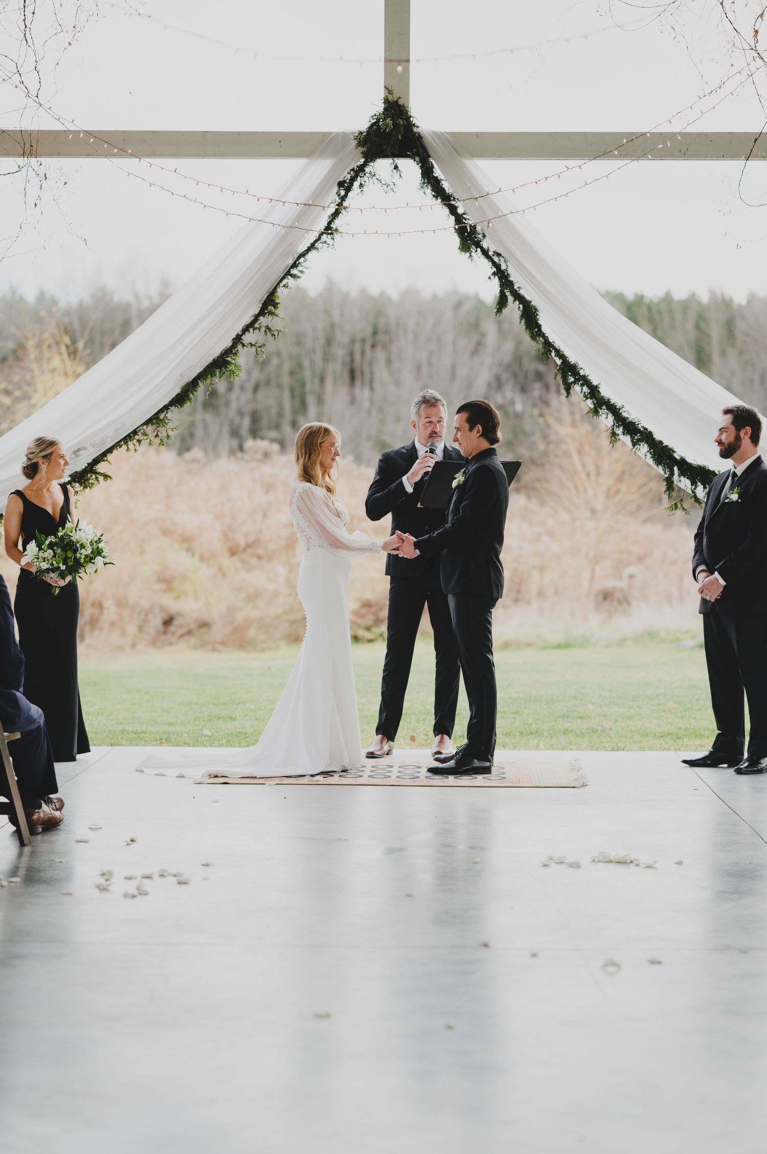 Wedding ceremony taking place under a white draped canopy with greenery decorations in an outdoor setting.