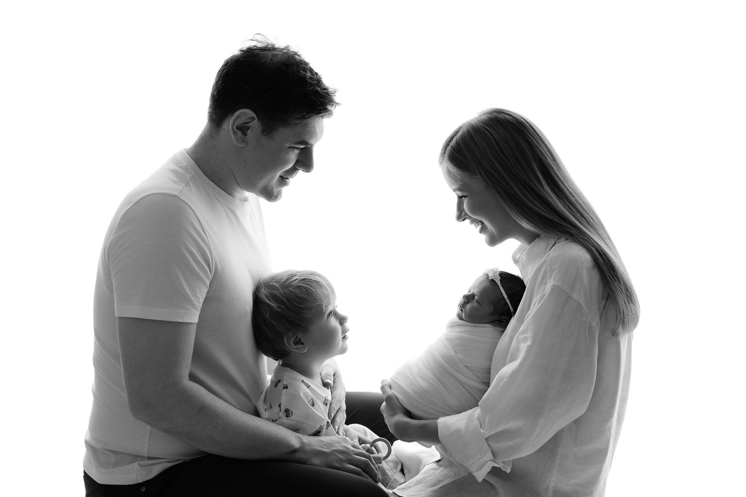 Black and white family portrait with parents lovingly holding a toddler and newborn baby against white background.