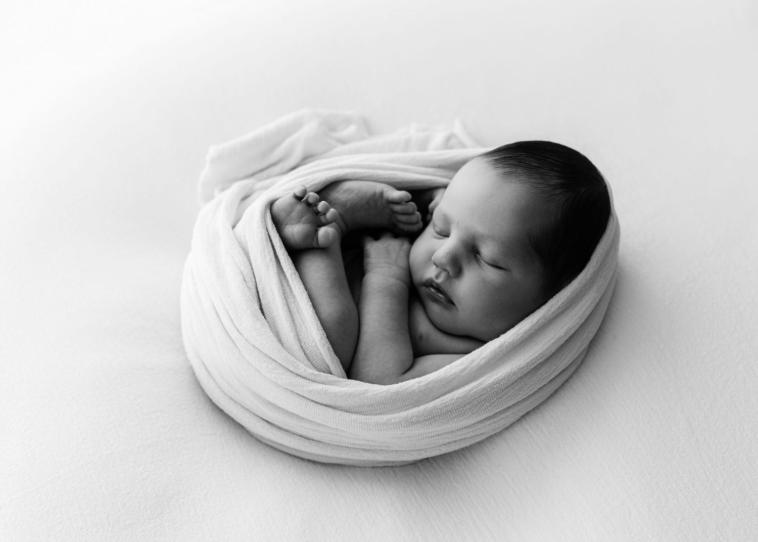 Black and white newborn baby wrapped in soft white cloth, sleeping peacefully in a cozy bundle pose.