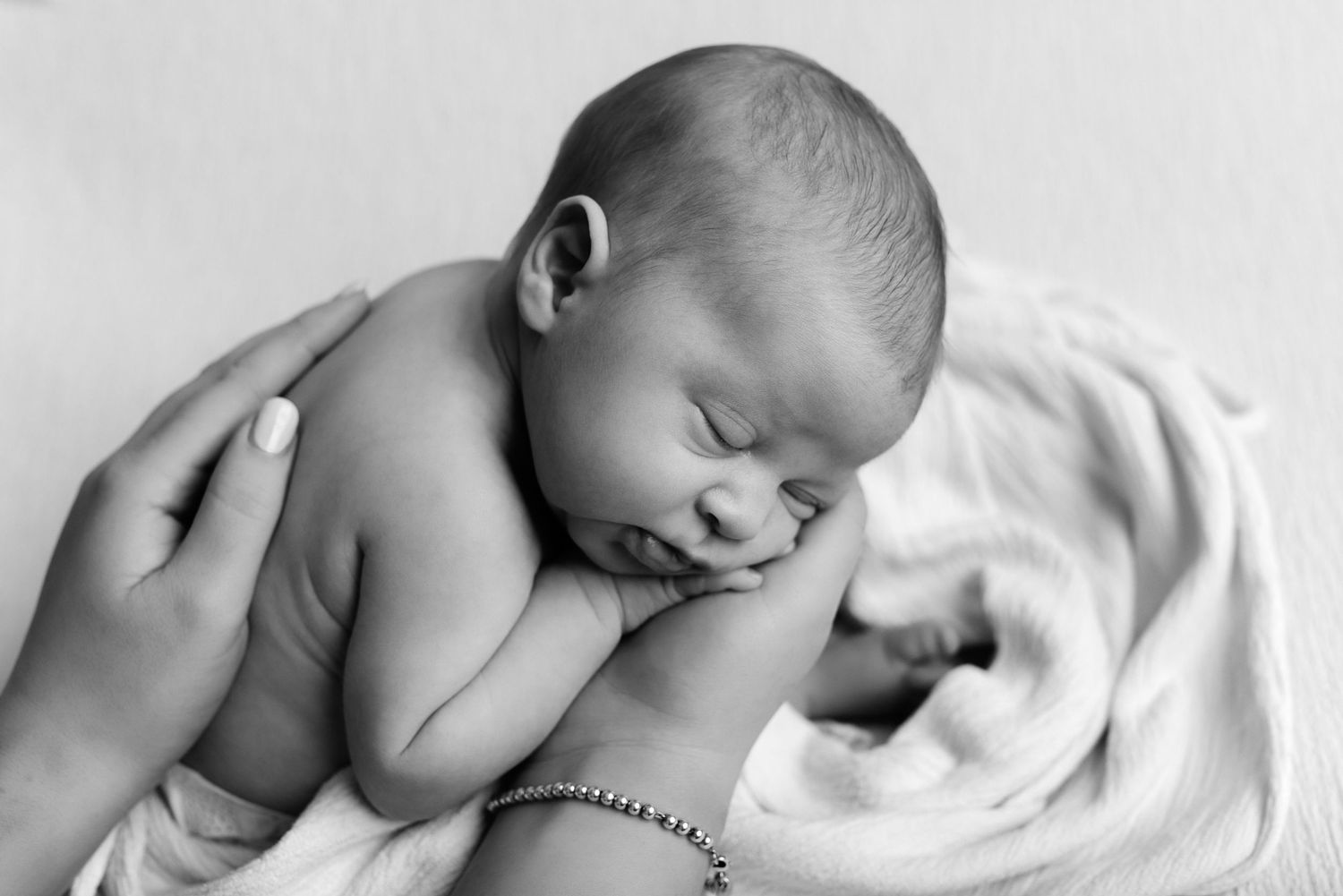 Black and white newborn baby sleeping peacefully in parent's hands on soft white blanket.
