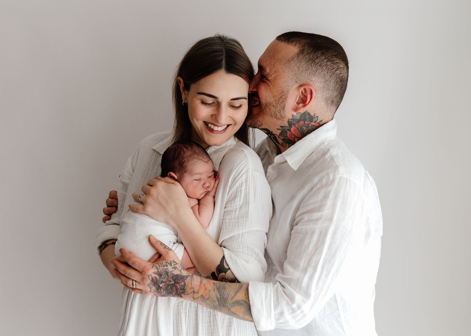 Smiling couple in white outfits tenderly holding their newborn baby, with the man kissing the woman's cheek.