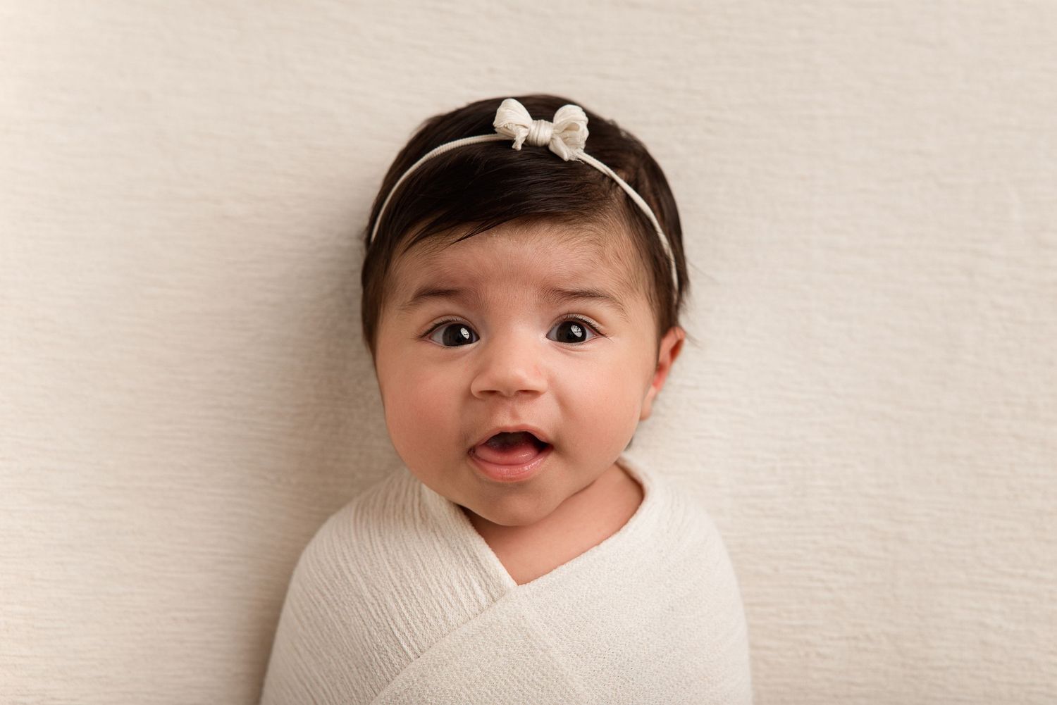 Adorable baby girl wrapped in white swaddle with bow headband, looking at camera against neutral background.